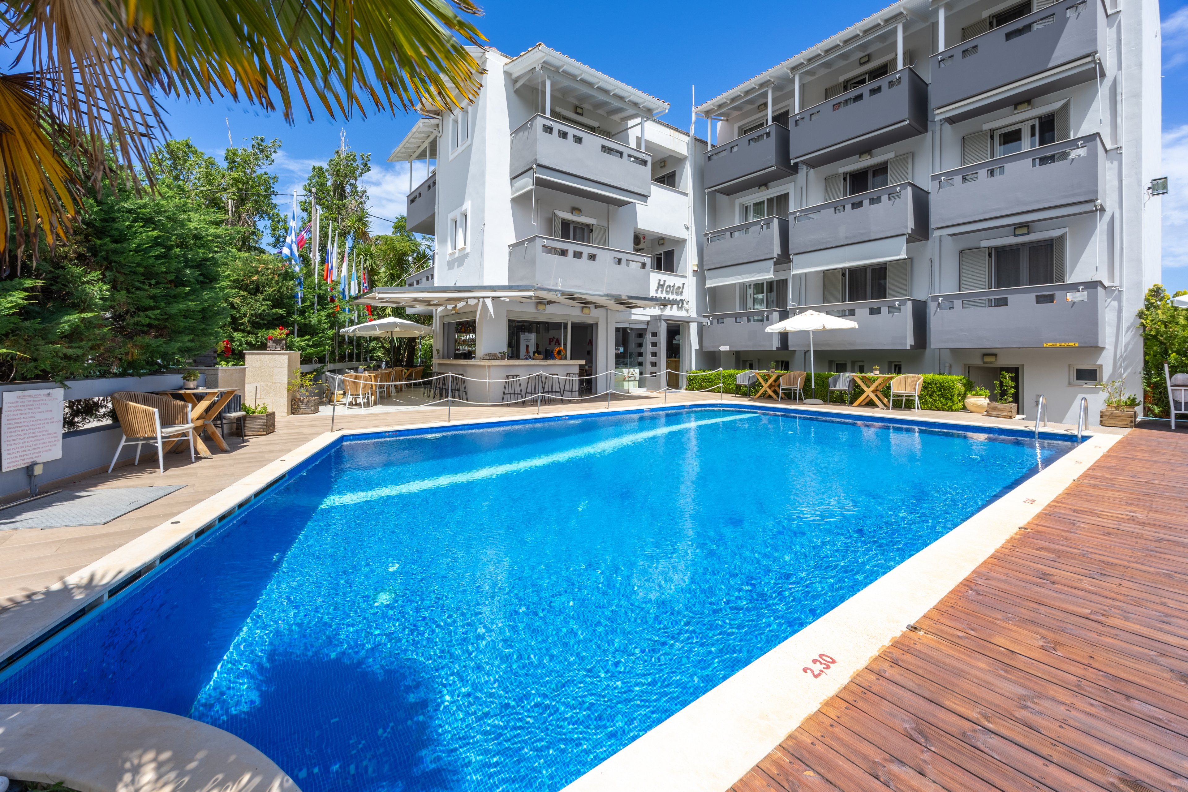 A bright blue outdoor swimming pool sits in front of a modern multi-story hotel building. Sun loungers and umbrellas line the poolside deck, creating a relaxing vacation atmosphere. The hotel features balconies, an open-air bar area, and surrounding greenery under a clear blue sky. The pool edge shows a depth marking of 2.05 m near the wooden deck.