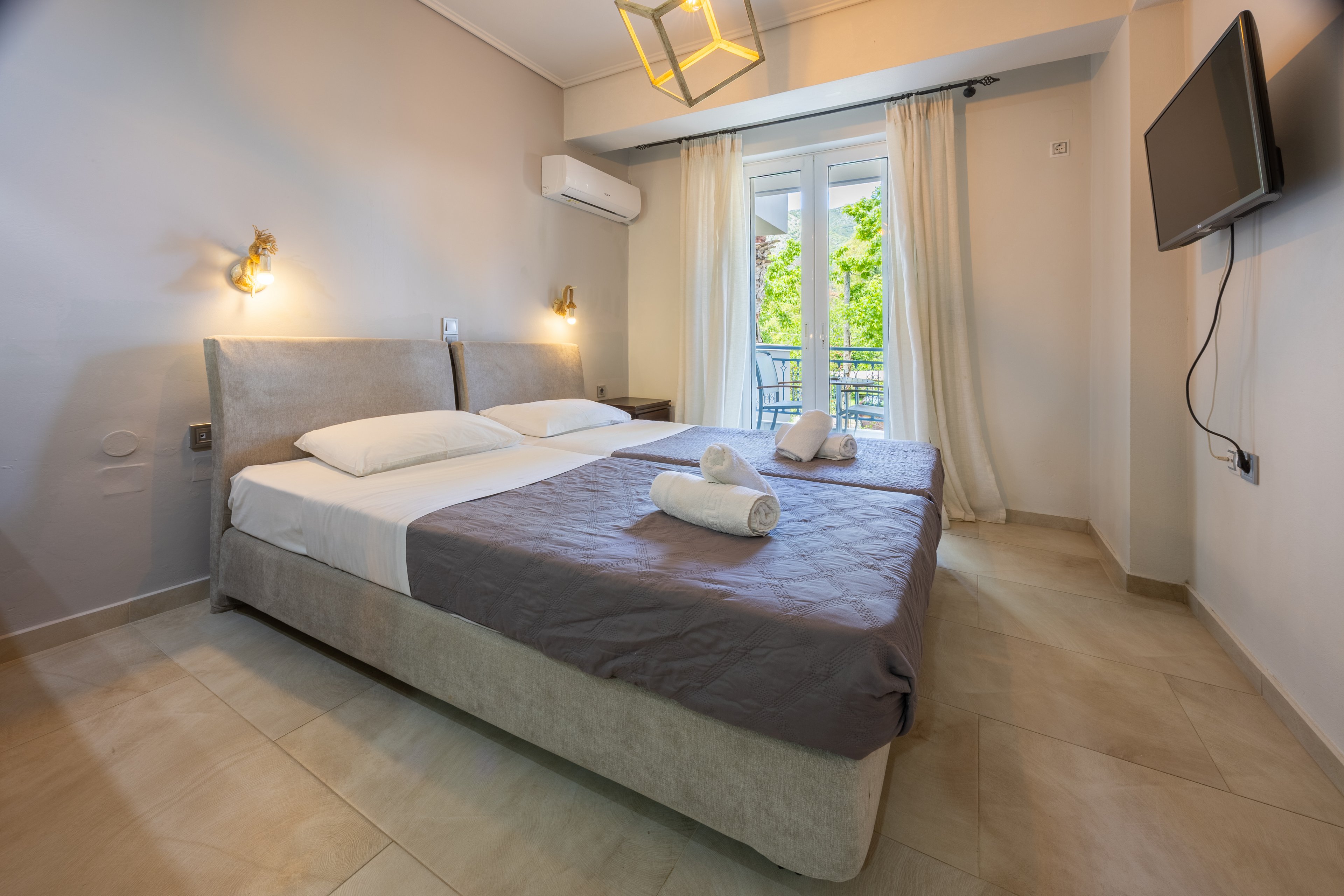 A modern hotel bedroom features two single beds pushed together with neatly folded white towels on top. The room has light neutral walls, tiled flooring, and minimalist wall-mounted lamps. A flat-screen television is mounted on the wall opposite the bed, and an air conditioning unit is installed above. Sheer curtains frame glass doors that open to a balcony with greenery visible outside, creating a bright and inviting atmosphere.