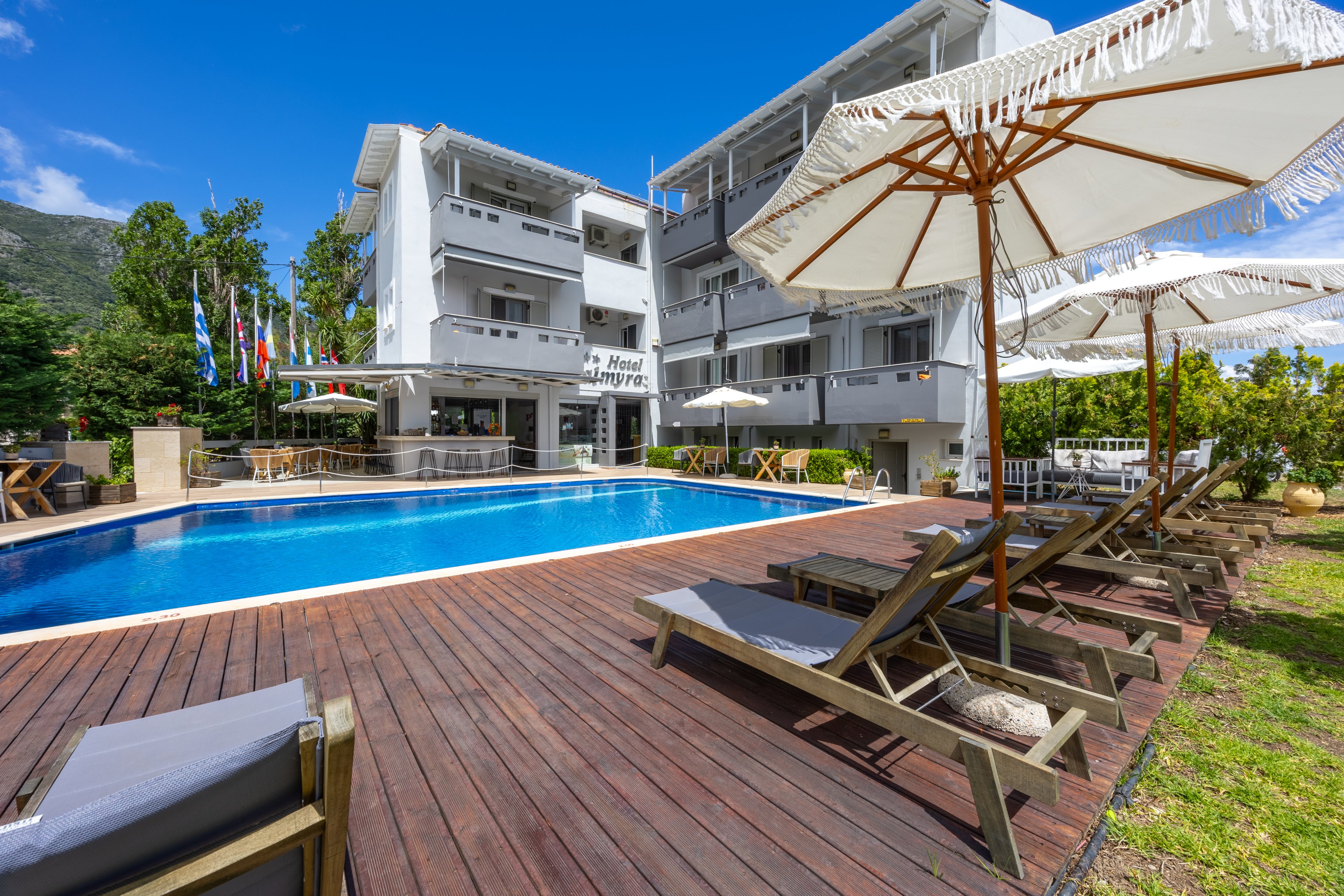 A contemporary multi-story hotel building overlooks a clear blue outdoor swimming pool. Wooden decking surrounds the pool, lined with sun loungers and large white parasols for guests to relax. The scene is set on a bright sunny day with a vivid blue sky and green trees in the background. Several national flags are visible near the hotel entrance, suggesting an international vacation destination.