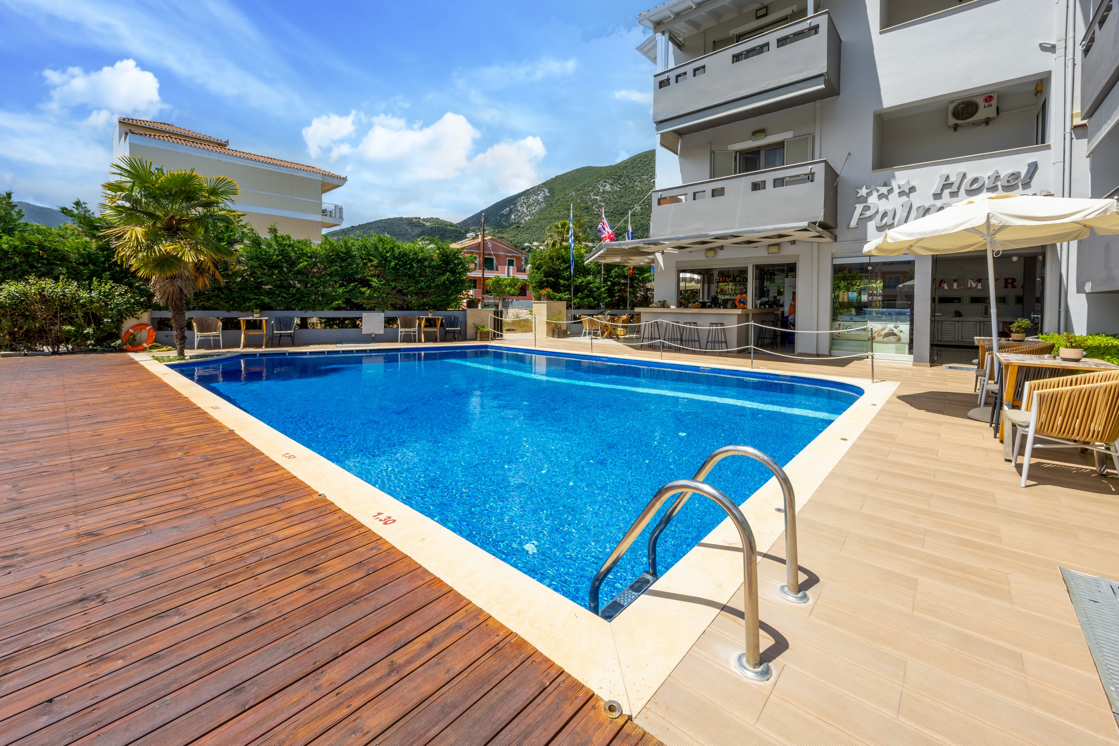 An inviting blue swimming pool sits in front of a modern hotel building on a bright sunny day. Wooden decking and tiled flooring surround the pool, with metal handrails leading into the water. Outdoor tables, chairs, and umbrellas create a relaxed seating area near the hotel bar or reception. Green shrubs and distant mountains frame the scene under a partly cloudy sky.