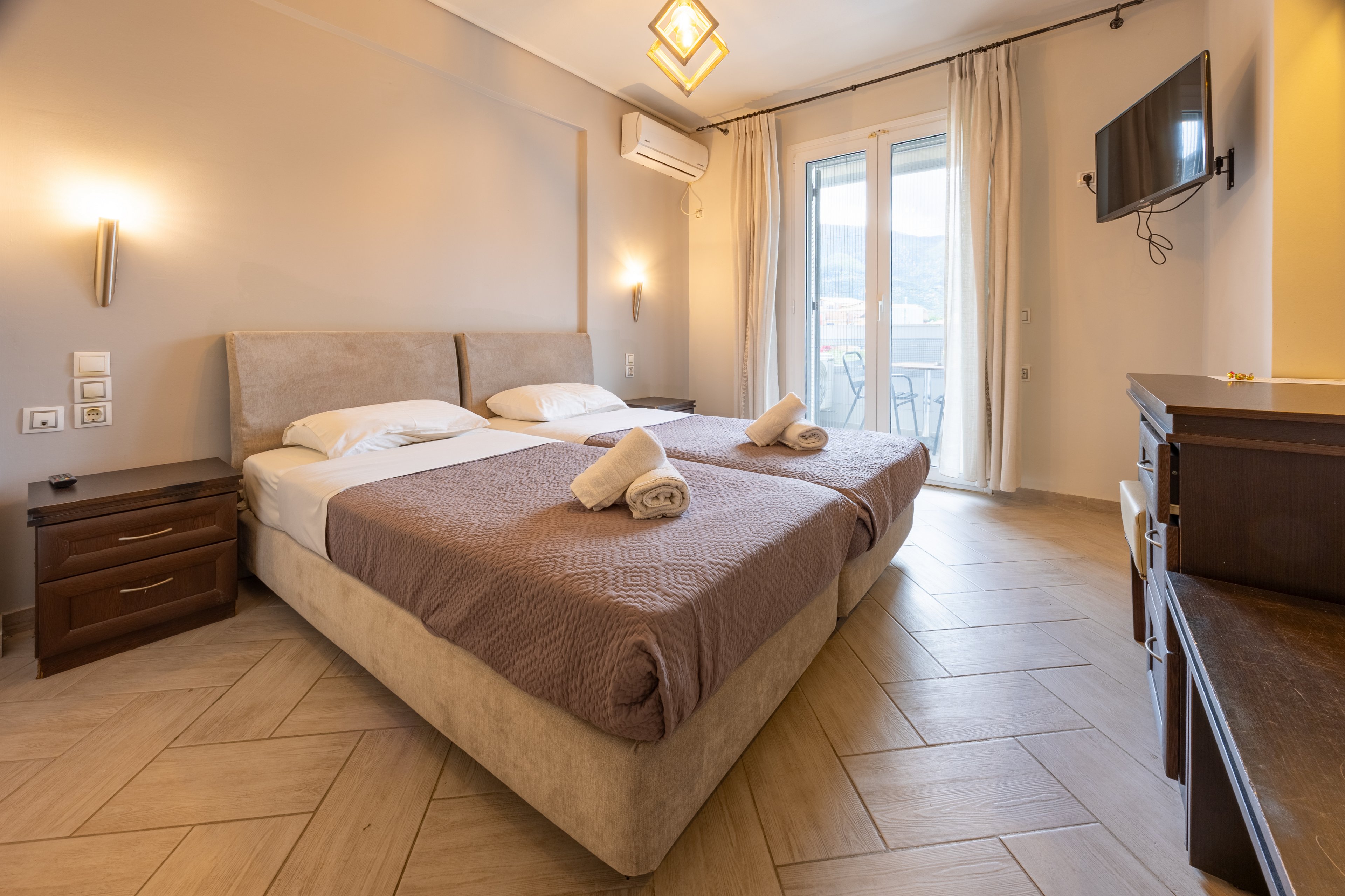 A warm, inviting hotel bedroom features two single beds pushed together with neatly folded towels on top. Soft beige and brown tones create a calm, neutral atmosphere. A wall-mounted television faces the bed, and a desk with a chair sits to the side. Large glass doors open to a balcony, allowing natural light to brighten the room.