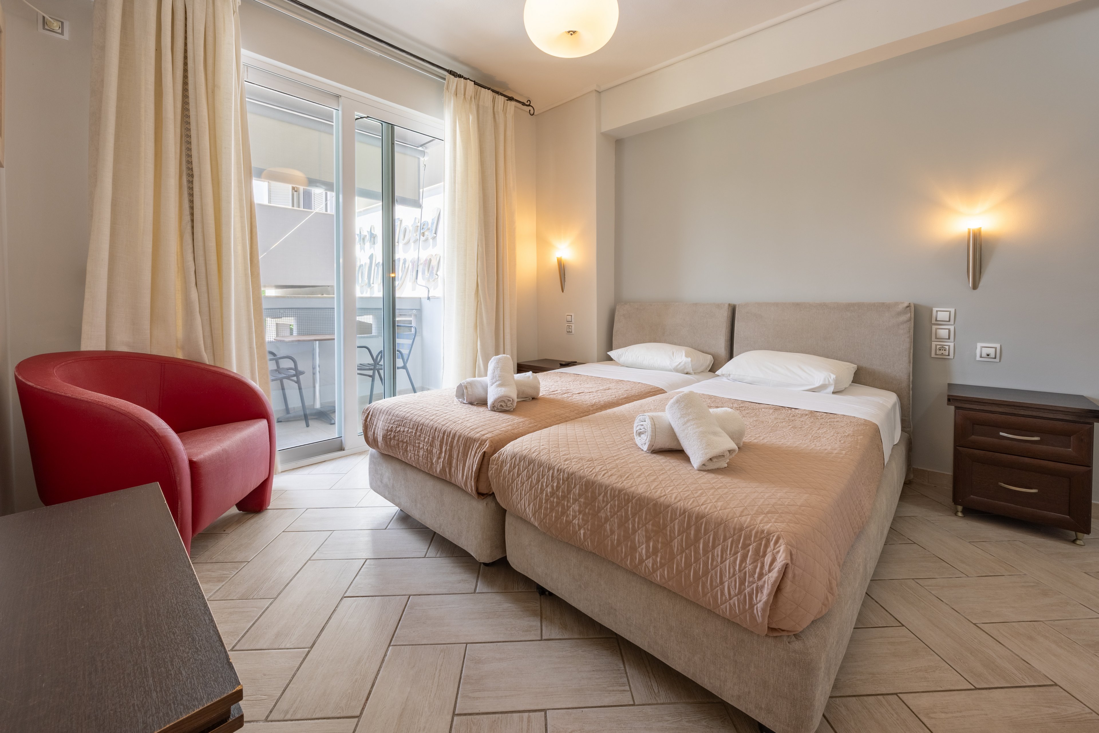 A modern hotel room features two neatly made twin beds with folded white towels placed on top. The space is bright, with natural light streaming in through large sliding glass doors that open to a small balcony with outdoor seating. Neutral beige and gray tones dominate the room, accented by a bold red armchair and warm wall lighting. The overall atmosphere is clean, inviting, and comfortable for guests.
