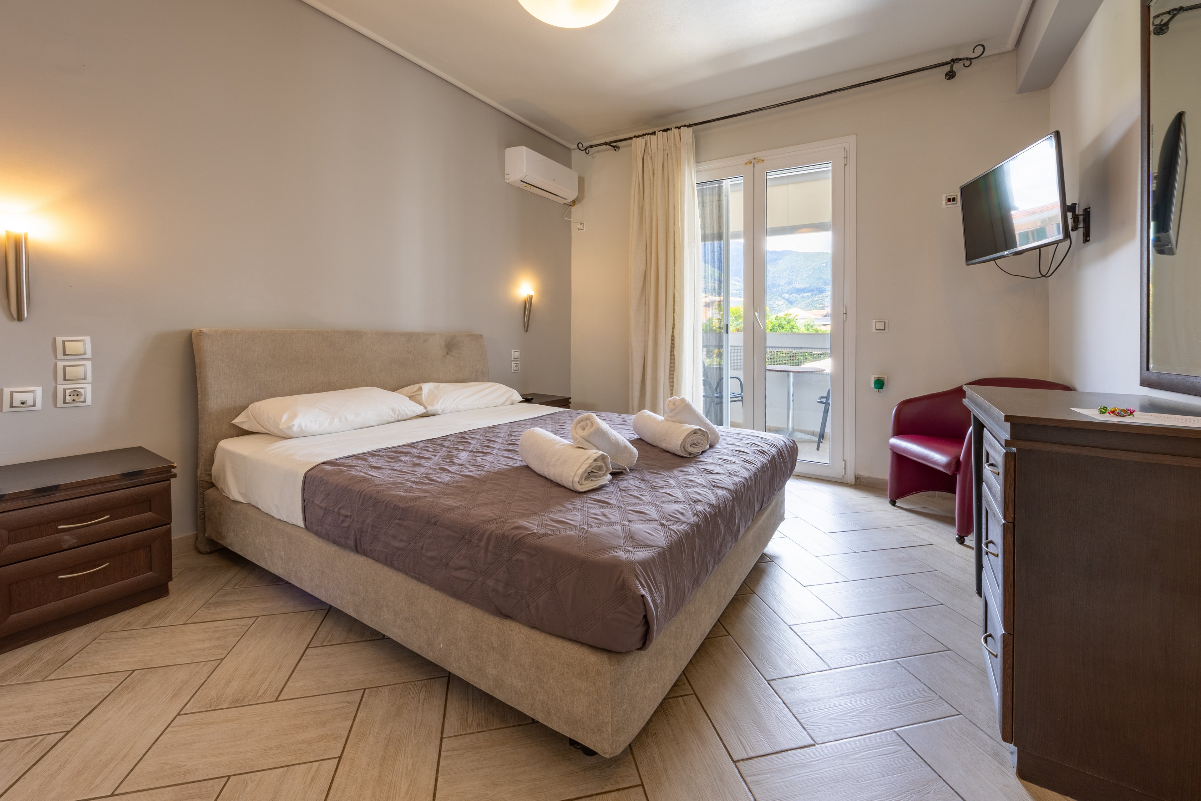 A neatly made double bed with folded towels is the focal point of this modern hotel bedroom. The room features neutral walls, wooden furniture, and a wall-mounted television opposite the bed. Large glass doors open to a balcony with a scenic mountain view, allowing natural light to fill the space. Warm wall sconces and tiled flooring contribute to the clean, inviting atmosphere.