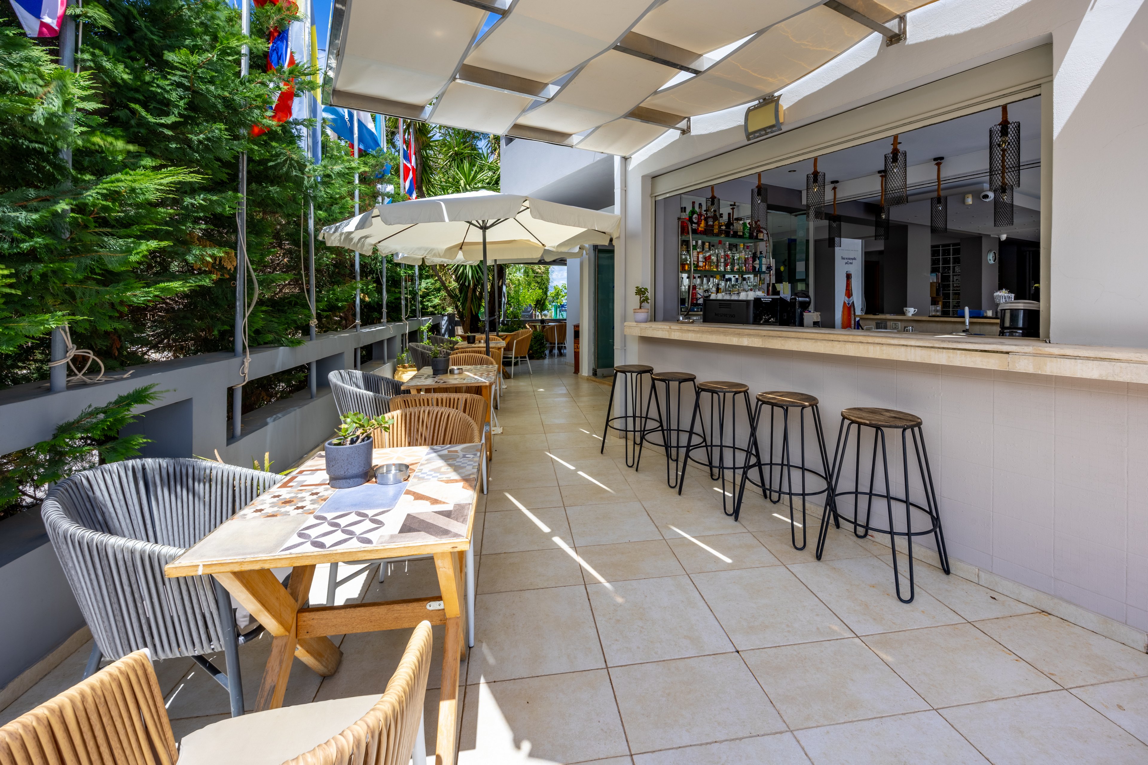 An open-air bar area features a long counter with several high metal stools lined up in front. A wooden table with woven chairs and a small potted plant sits on the tiled floor, creating a cozy seating corner. Large umbrellas provide shade, while lush green trees and international flags add color along the side. The bright daylight and clean, modern design give the space a relaxed vacation atmosphere.