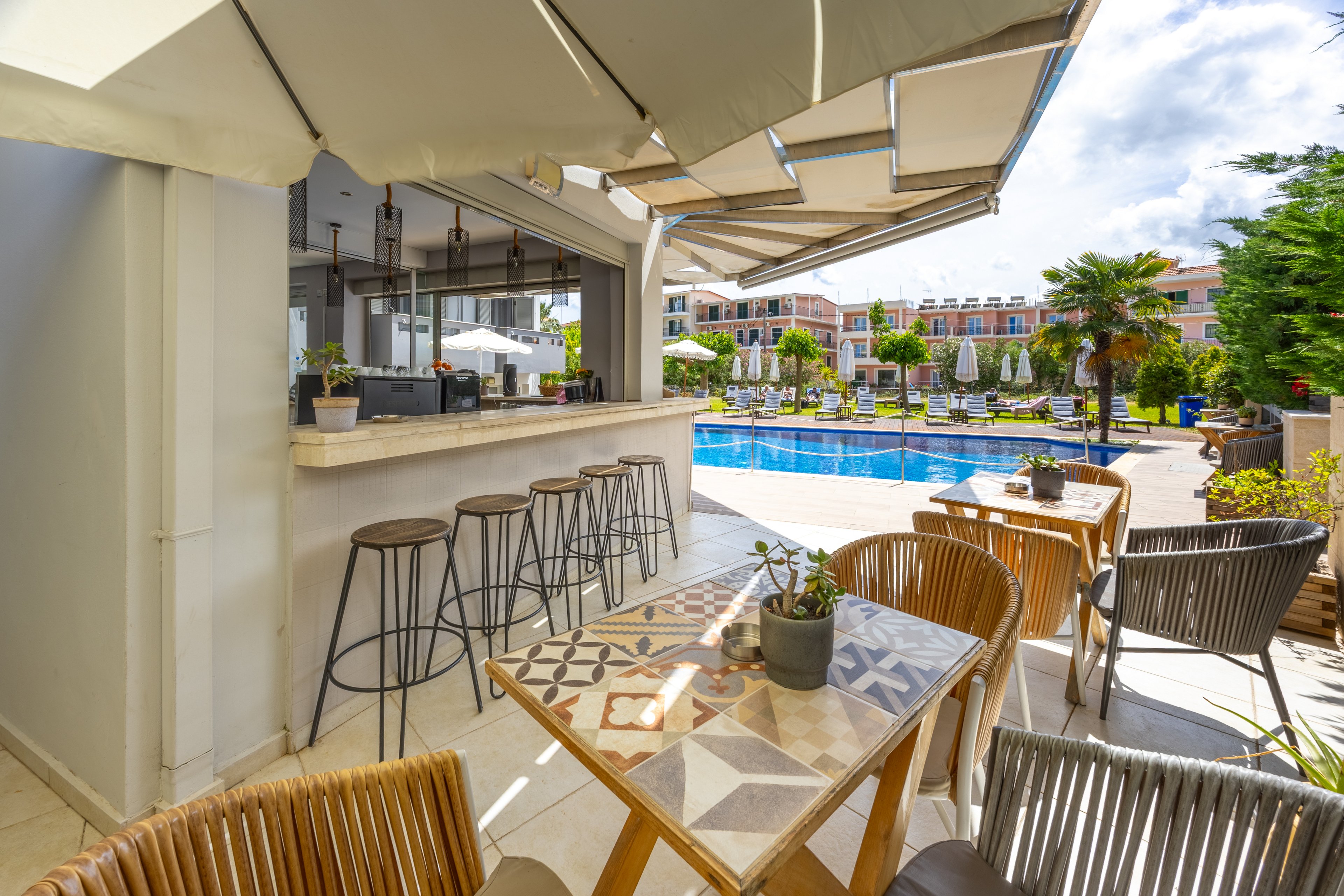 An inviting outdoor bar area overlooks a bright blue swimming pool in a resort setting. High metal bar stools line the counter under a large awning, while wooden tables and woven chairs provide relaxed seating in the foreground. Potted plants and patterned tabletops add decorative detail to the scene. In the background, sun loungers, umbrellas, and low-rise buildings suggest a sunny vacation destination.