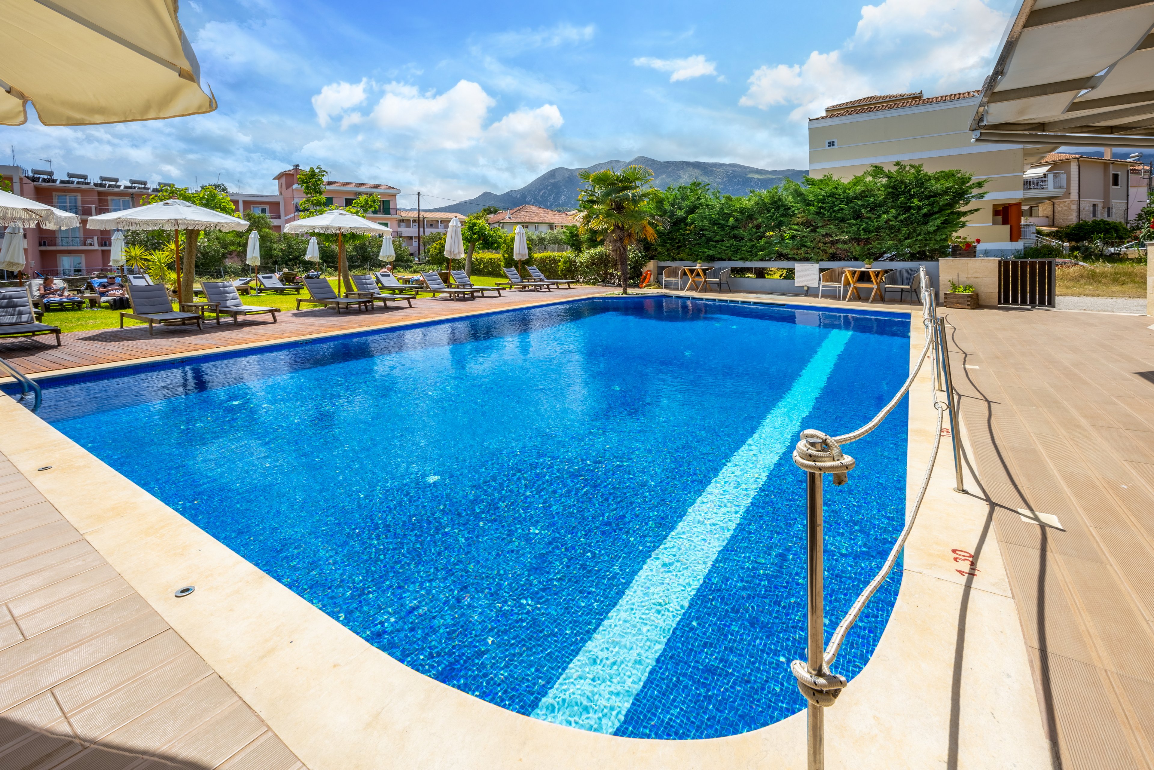 A clear blue outdoor swimming pool is the central focus of this resort scene. Sun loungers and umbrellas line the poolside deck, suggesting a relaxing vacation setting. Residential-style buildings and manicured greenery surround the area, with mountains visible in the distant background. Bright daylight and a vivid blue sky create a warm, inviting atmosphere for leisure and sunbathing.