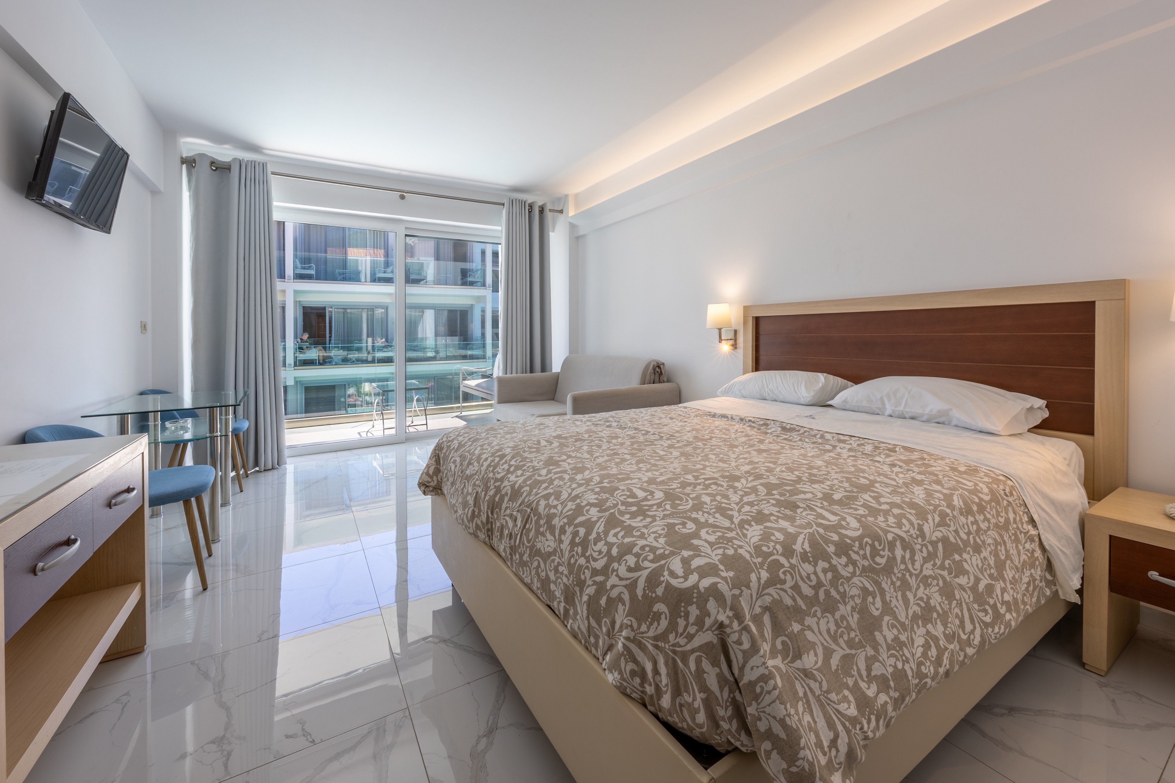 A spacious modern hotel bedroom features a large double bed with a patterned beige duvet. The room includes a small glass dining table with two chairs, a wall-mounted television, and a light-colored desk. Floor-to-ceiling windows with sheer and blackout curtains open onto a balcony overlooking neighboring buildings. Neutral tones and glossy tile flooring create a clean, minimalist, and inviting atmosphere.