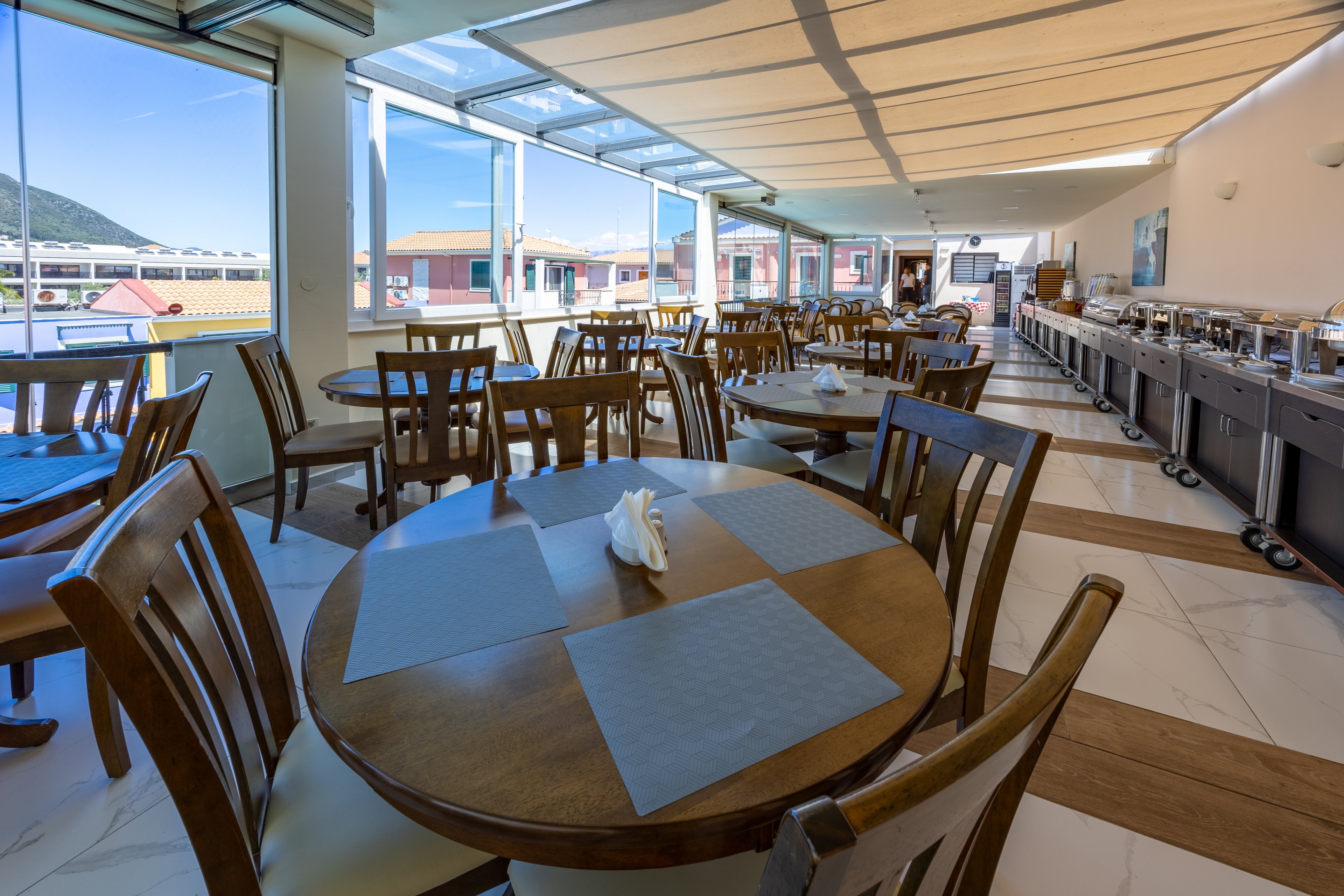 A spacious hotel restaurant dining area features wooden tables set with placemats and folded napkins. Large floor-to-ceiling windows provide abundant natural light and views of nearby buildings and distant hills. Along one wall, a long buffet counter is prepared for self-service meals with covered dishes and serving equipment. The clean, modern interior has tiled floors, neutral colors, and a partially covered ceiling creating a relaxed, airy atmosphere.