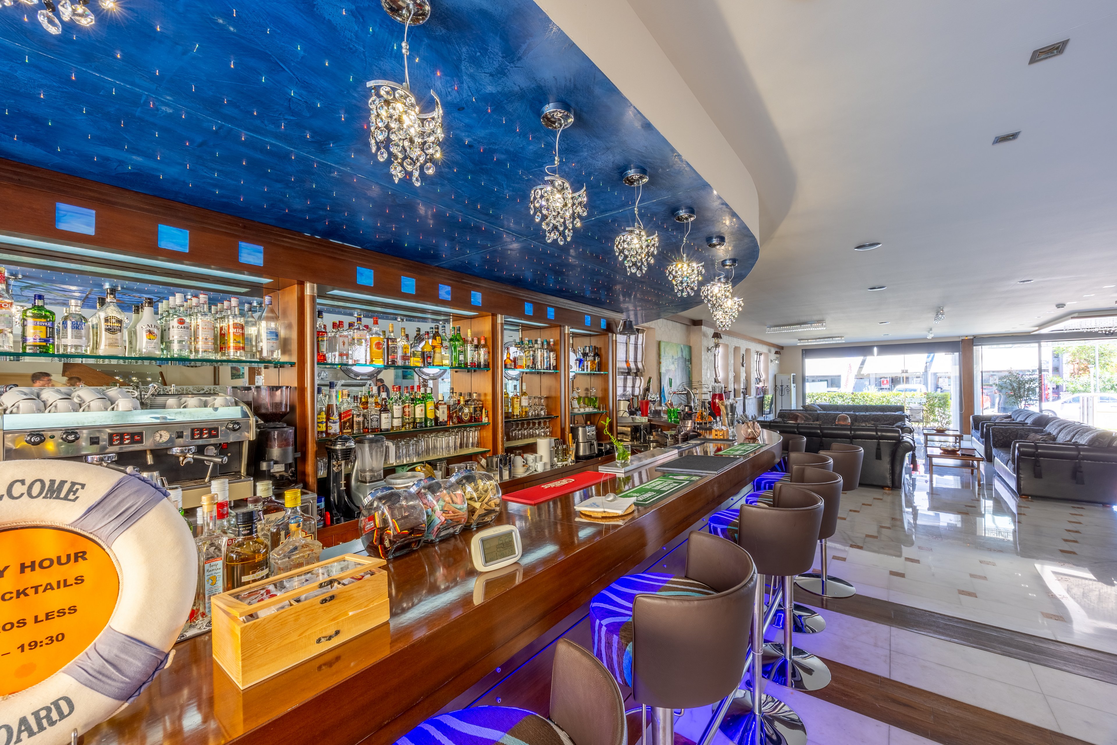A contemporary bar interior features a long counter lined with modern bar stools and a fully stocked back bar of spirits and glassware. The ceiling above the bar is painted a vivid blue and accented with sparkling pendant lights, creating a stylish focal point. Large windows in the background let in natural light and reveal additional lounge seating areas. The overall scene suggests a relaxed yet upscale atmosphere suitable for socializing or enjoying cocktails.
