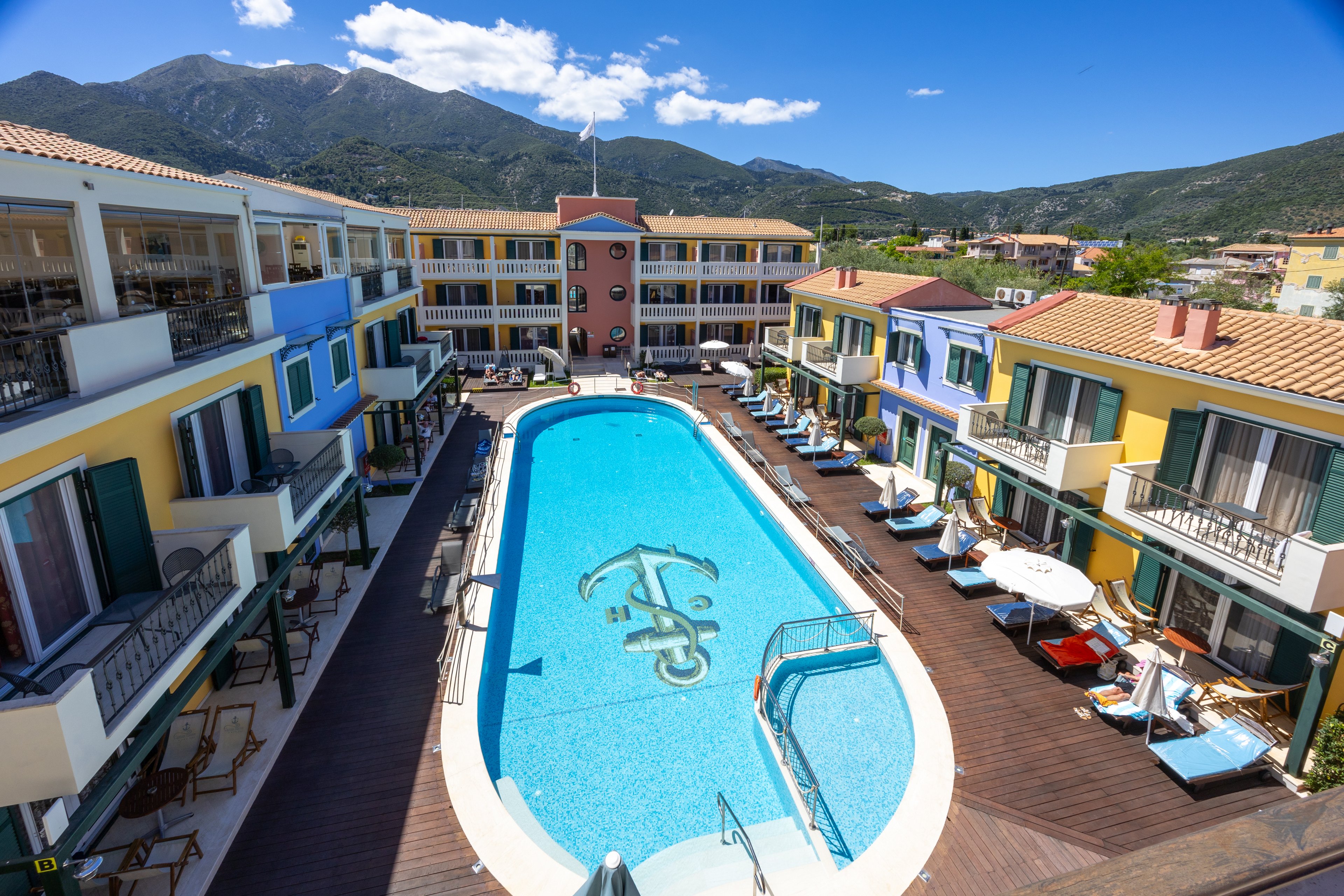 A bright resort complex surrounds a long, narrow swimming pool decorated with an anchor design on the pool floor. Colorful multi-story buildings with balconies frame the wooden deck area, where sun loungers and umbrellas line both sides of the pool. The scene is set against a backdrop of green mountains and a blue sky with scattered clouds. The overall atmosphere suggests a sunny, relaxing vacation destination.