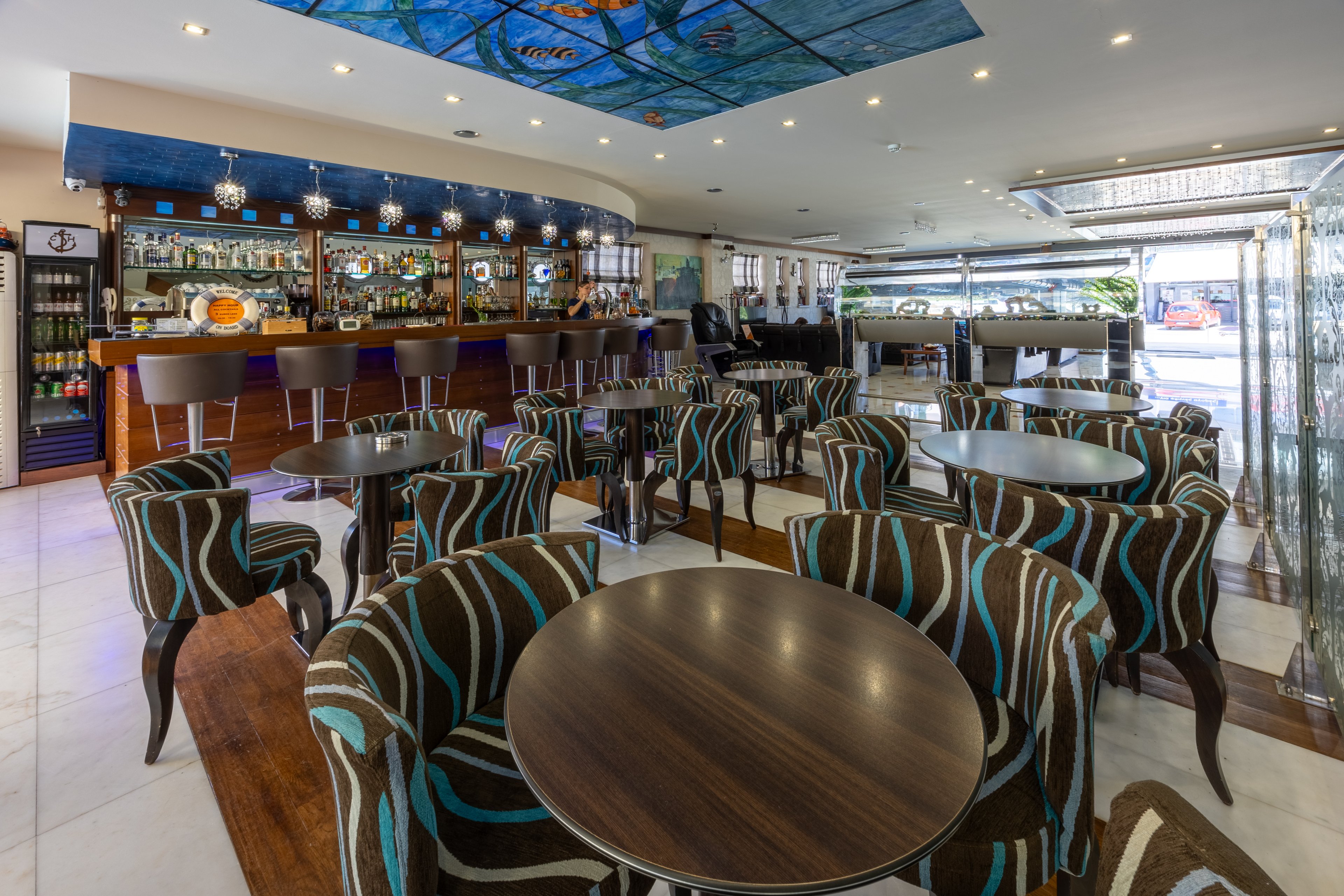 A contemporary café bar interior features multiple round wooden tables surrounded by upholstered chairs with bold blue and brown patterns. The bar counter at the back is stocked with bottles, glassware, and refrigerated displays, with high stools lined along the front. Large windows and an open entrance bring in natural light and a view of the street outside. Recessed ceiling lights and a decorative blue ceiling panel add to the modern, inviting atmosphere.