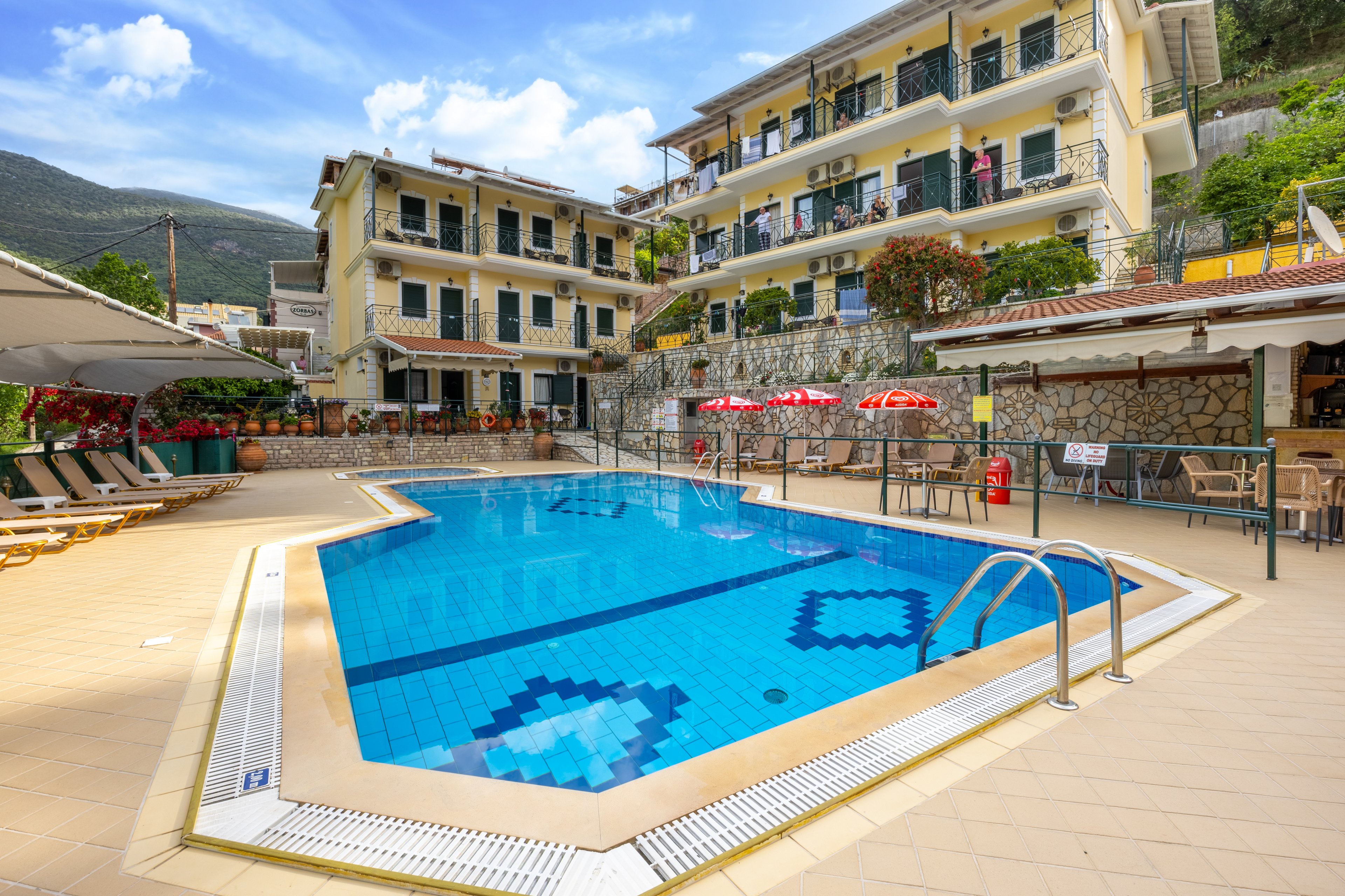 An outdoor swimming pool sits at the center of a sunny hotel courtyard. Multi-story buildings with balconies and green shutters surround the pool area, backed by tree-covered hills. Lounge chairs and umbrellas line the tiled deck, with a small bar or café area visible to the side. The clear blue water and bright sky create a relaxed vacation atmosphere.