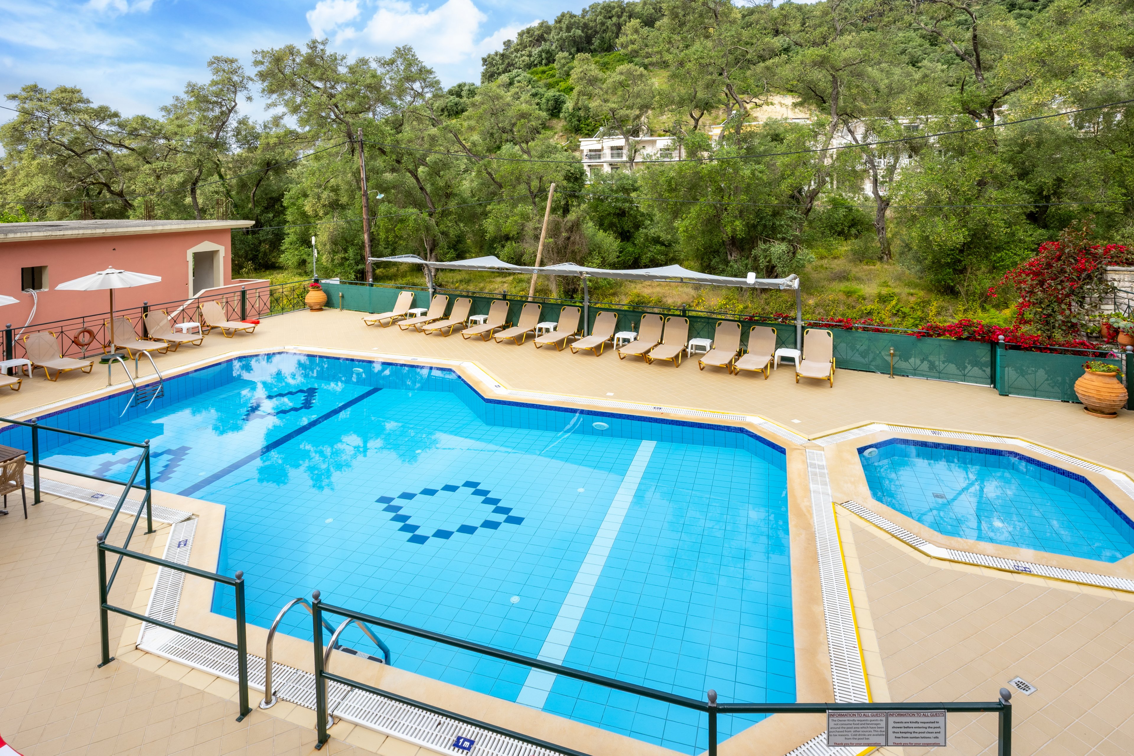 An outdoor hotel swimming pool is surrounded by numerous empty sun loungers and parasols. The clear blue water features a decorative tile pattern in the center and a shallow entry area with metal handrails. A smaller adjacent pool sits to the side, bordered by beige tiles and safety fencing. Lush green trees, flowering shrubs, and nearby buildings create a tranquil resort atmosphere under a bright sky.