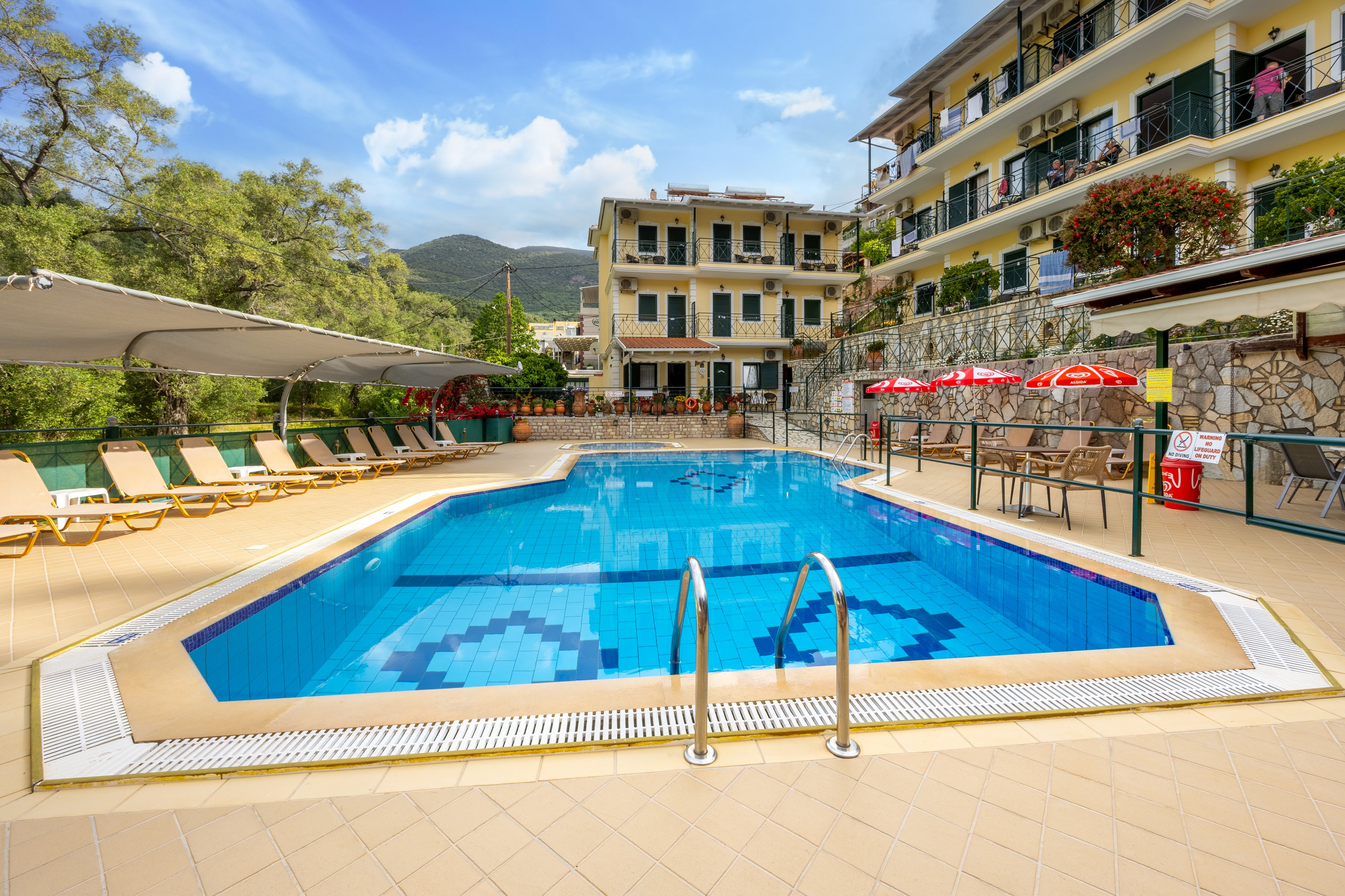 An outdoor swimming pool sits at the center of a sunny hotel courtyard. Lounge chairs line the tiled deck under shade canopies, with red umbrellas and a small bar area on one side. Multi-story buildings with balconies and flowering plants surround the pool, and green hills rise in the background under a blue sky. The scene suggests a relaxed vacation atmosphere at a resort or guesthouse.