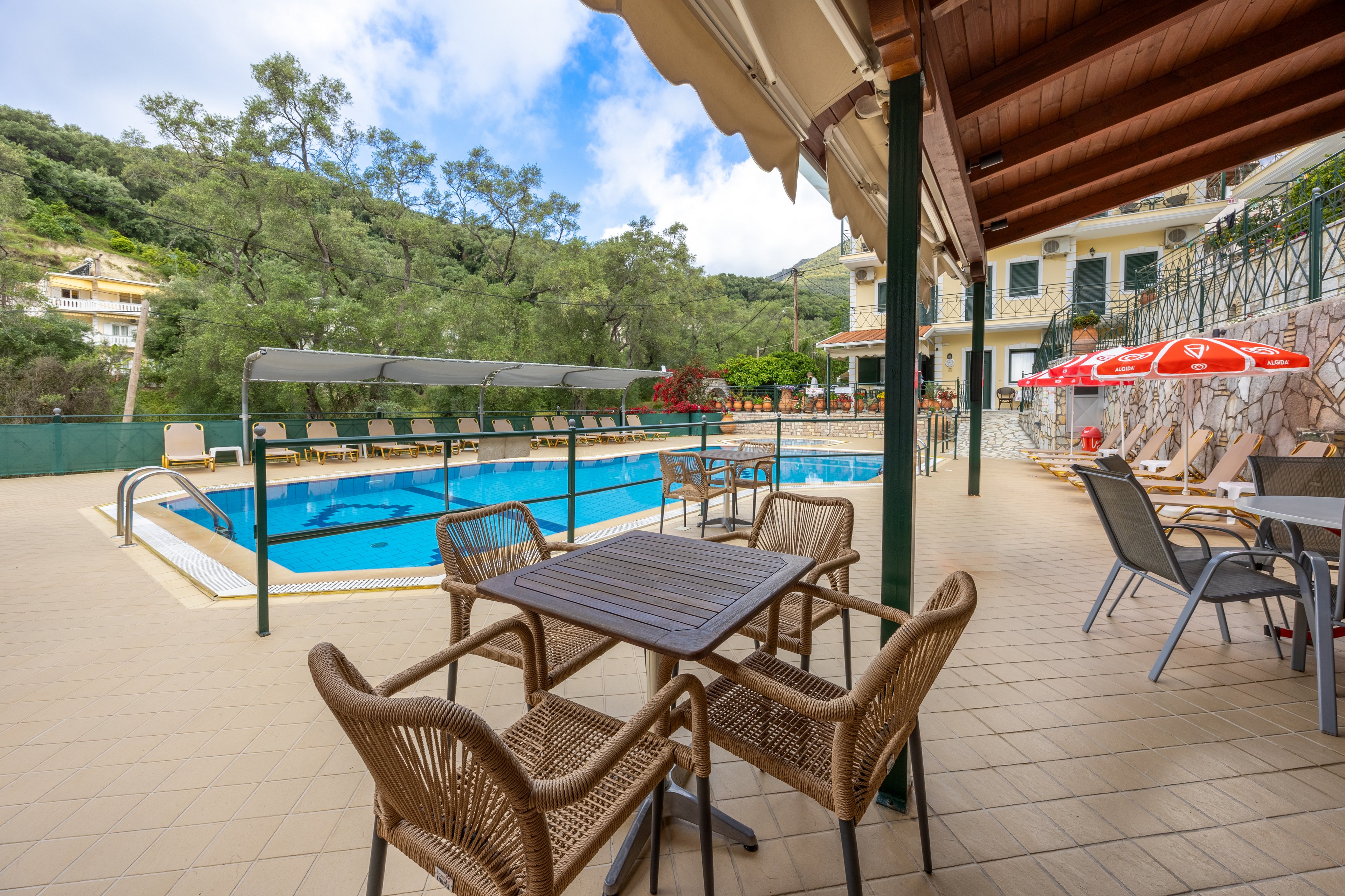 An outdoor hotel swimming pool is surrounded by lounge chairs and a tiled terrace. In the foreground, wicker chairs and tables provide a shaded seating area under an awning. Red umbrellas and additional tables line the side of the pool near the building. Lush green hills and trees form a scenic natural backdrop behind the property. The scene suggests a quiet, relaxing vacation setting on a clear day.