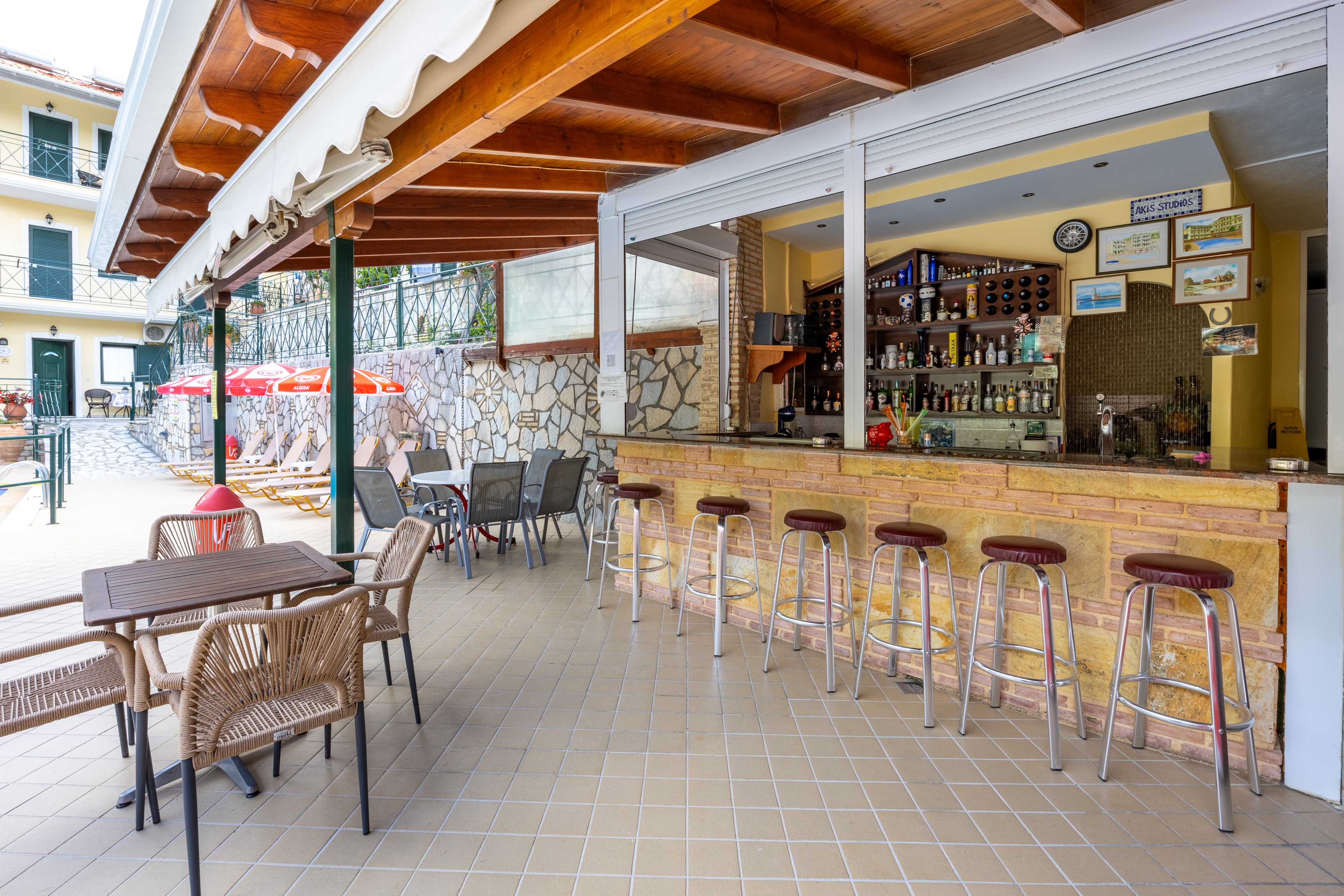 An open-air bar with a tiled floor features a long counter lined with metal bar stools. The bar shelves are stocked with various bottles and glasses, and framed pictures and a clock hang on the wall. In the foreground, wicker chairs and tables provide additional seating under a wooden awning. Red umbrellas and lounge chairs are visible outside, suggesting a poolside or resort setting on a bright day.