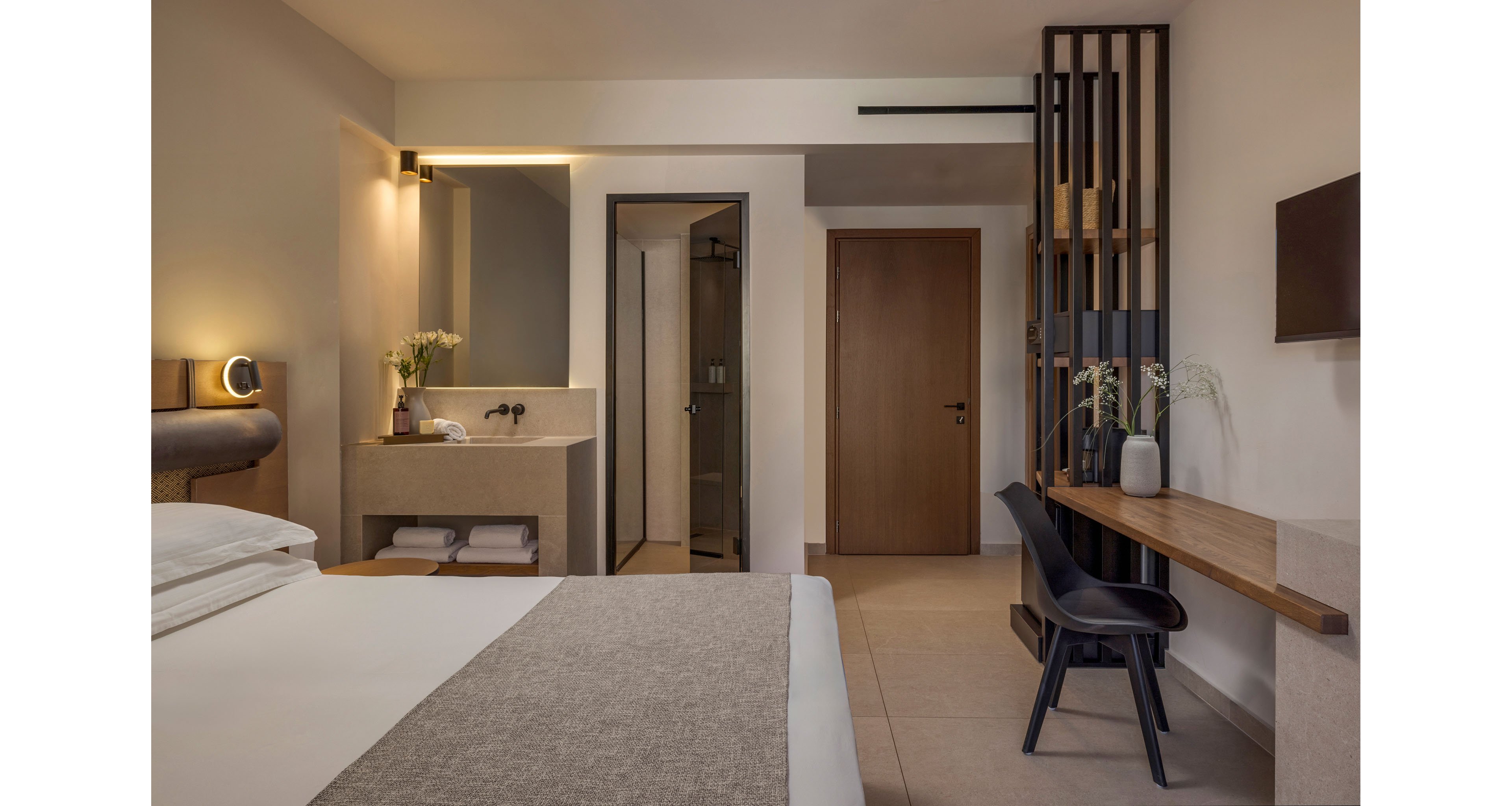 A contemporary hotel room features a neatly made bed in the foreground and a compact vanity area with a sink and flowers to the left. A wooden door and open doorway lead to the bathroom, adding depth to the space. On the right, a simple wooden desk with a dark chair and decorative vase creates a functional workspace beneath a wall-mounted television. Neutral tones, soft lighting, and clean lines convey a calm, minimalist atmosphere.