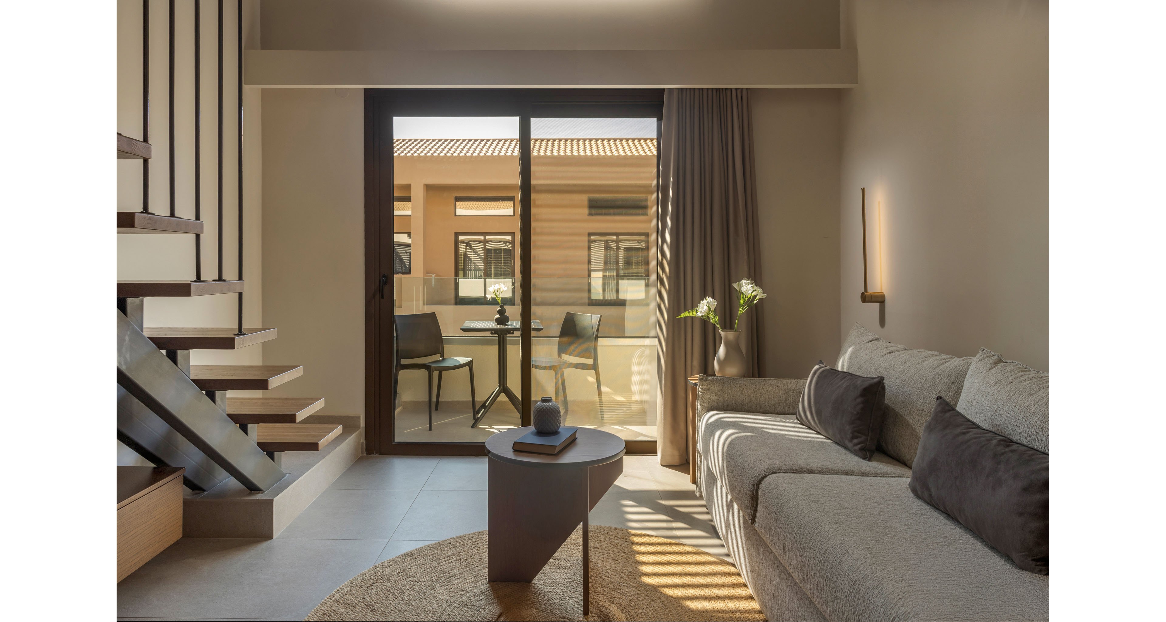 Contemporary living room interior features a light-colored sofa with dark accent cushions and a minimalist round coffee table. A sliding glass door opens to a small balcony with two chairs and a table, overlooking neighboring buildings. Soft natural light fills the space, highlighting the staircase with wooden steps and the neutral-toned rug. Warm wall lighting and a potted plant add a cozy, inviting atmosphere to the modern design.