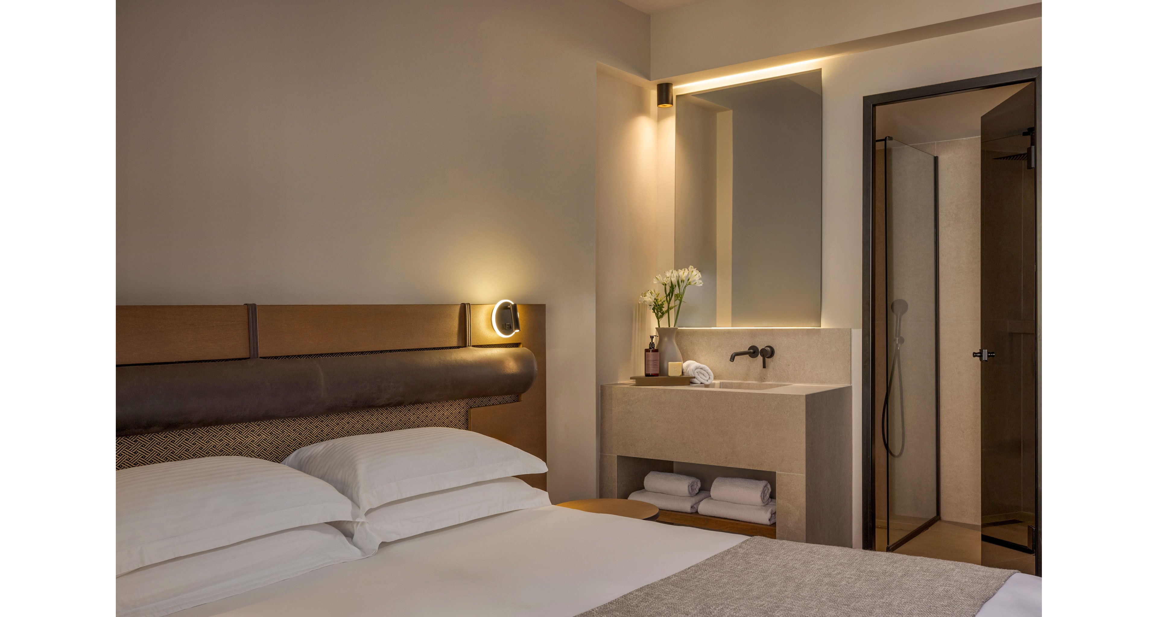 A contemporary hotel room features a neatly made bed with crisp white pillows and linens. Beside the bed, a small vanity area includes a minimalist sink, faucet, and a vase of white flowers. Rolled towels are stored on the shelf below the sink, adding to the organized and clean look. In the background, an open doorway reveals a simple walk-in shower, all illuminated by warm, indirect lighting.