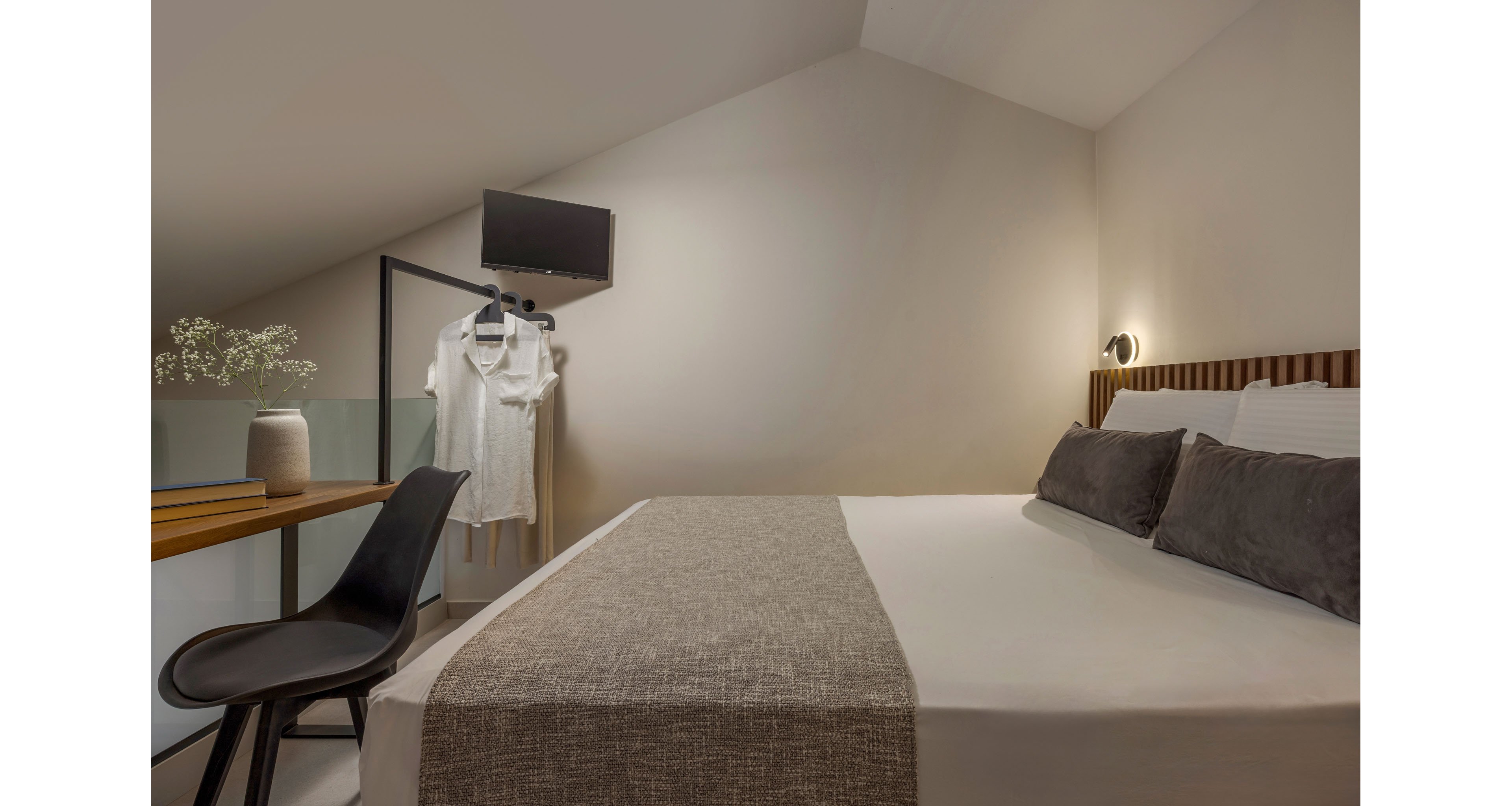 A cozy attic bedroom features a neatly made bed with neutral bedding and two dark accent pillows. A small desk with a black chair sits beside the bed, holding a vase with delicate white flowers. Opposite the bed, a simple clothing rack displays a white shirt beneath a wall-mounted television. Soft, warm lighting and sloped ceilings create an intimate, minimalist atmosphere.