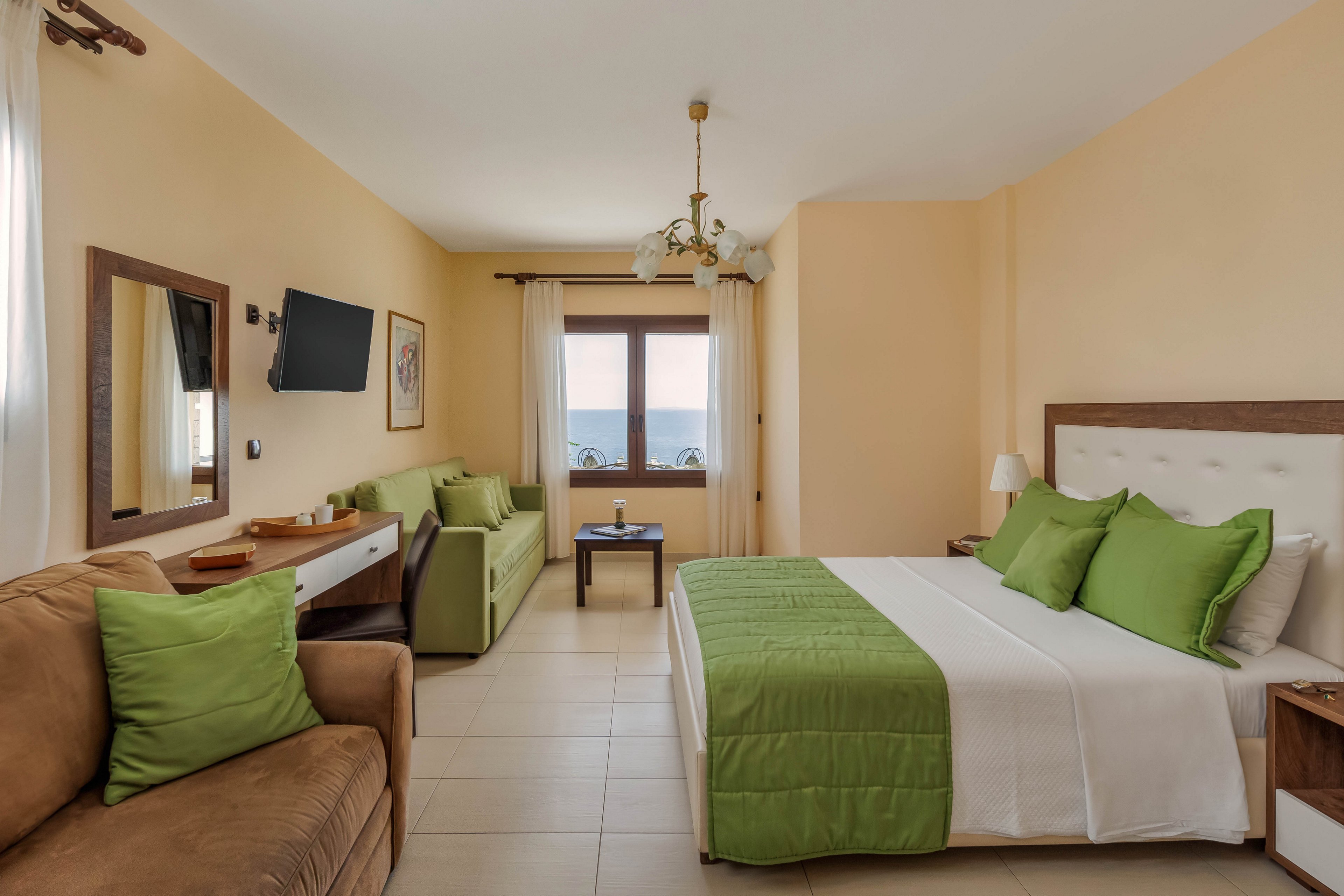 A spacious hotel bedroom features a large double bed with white linens and green pillows and bedspread. The room includes a brown sofa and a green couch along one wall, facing a wall-mounted television and a wooden-framed mirror. Natural light enters through a central window with curtains, offering a view of the sea in the distance. Neutral beige walls and tiled flooring create a clean, relaxing atmosphere complemented by simple decorative elements and a ceiling light fixture.