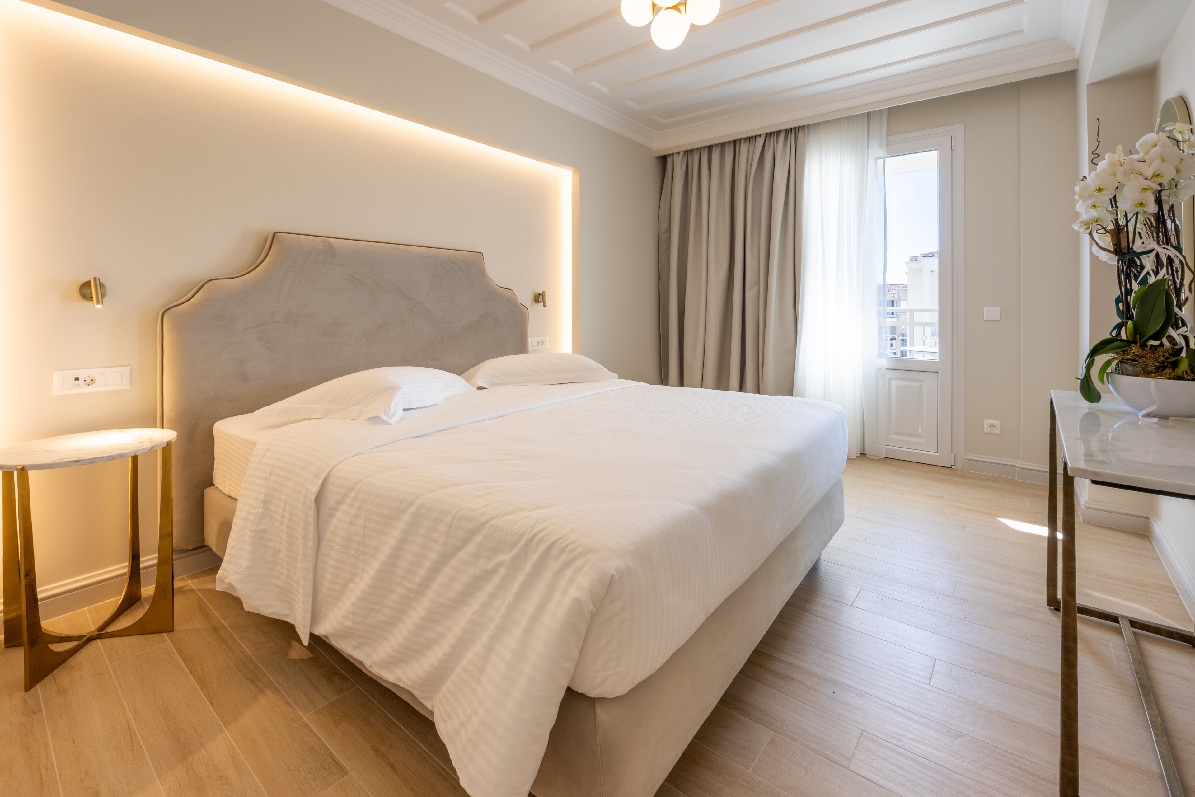 A neatly made double bed with white linens is the focal point of this modern bedroom. The room features a soft upholstered headboard with warm backlighting and light wood flooring. A small round side table and a console with flowers add simple decorative touches. Natural light enters through a tall window with sheer and blackout curtains, creating an inviting and serene atmosphere.