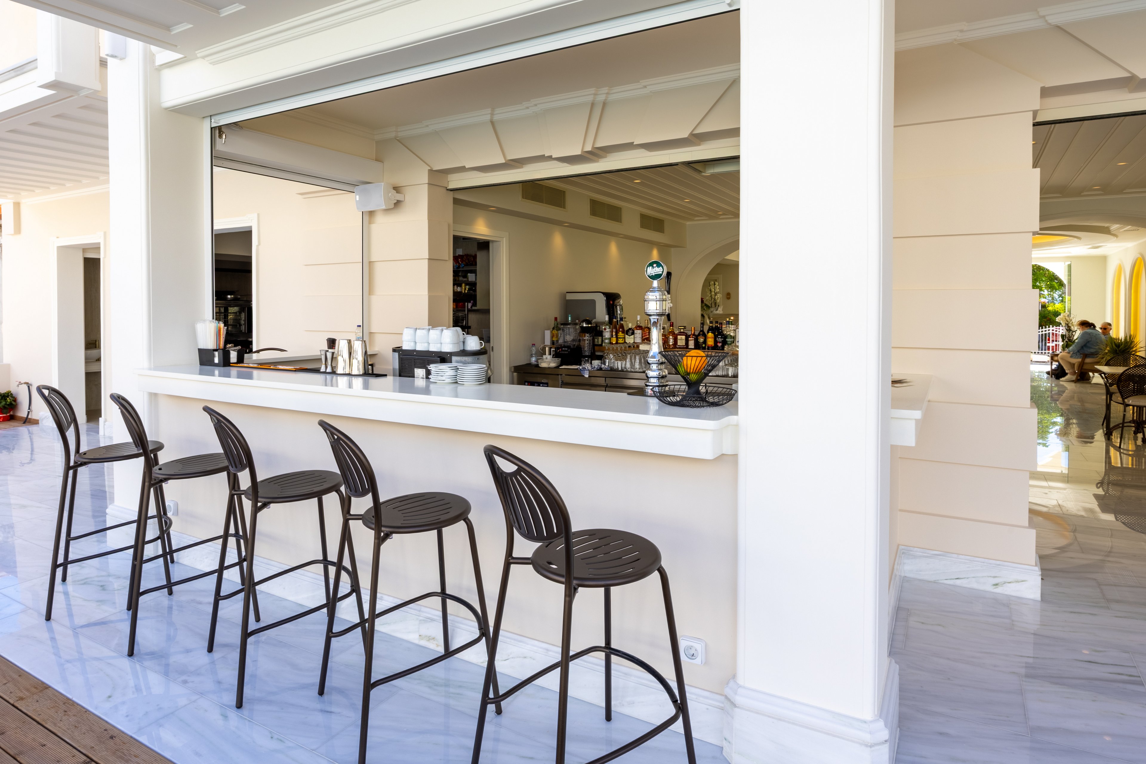 An open-air bar with a white counter and several tall metal stools stands ready for guests. The bar is integrated into a light-colored resort building with a wide serving window. Inside, bottles, glasses, and coffee equipment are neatly arranged on shelves and the back counter. In the background, a seating area with tables and chairs is visible, suggesting a relaxed vacation atmosphere.