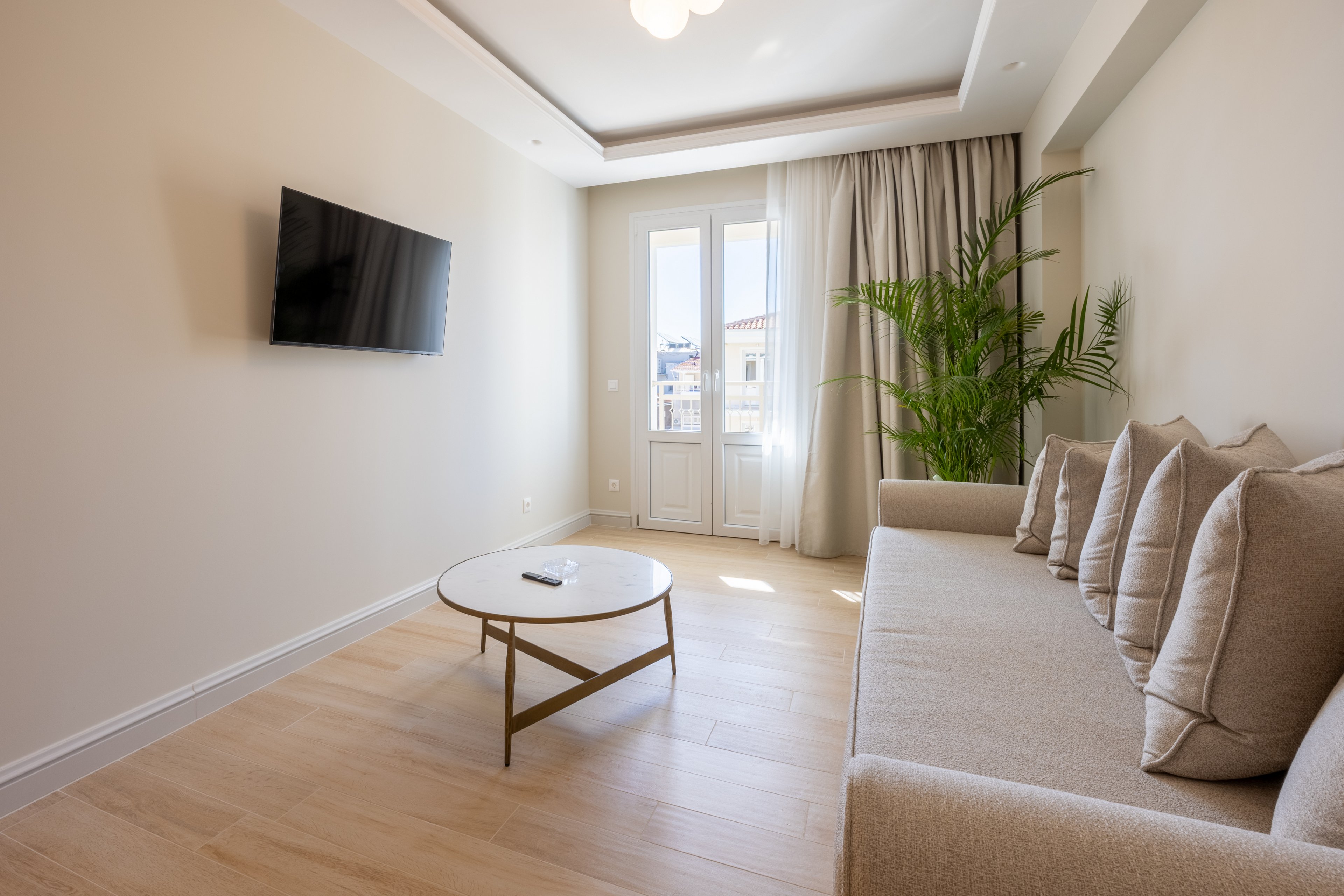 A bright minimalist living room features a long beige sofa with multiple cushions and a round white coffee table. A flat-screen television is mounted on the plain wall opposite the seating area. Natural light streams in through tall glass doors leading to a small balcony with a city view. Light wood flooring and a large green potted plant add warmth and freshness to the neutral-toned space.