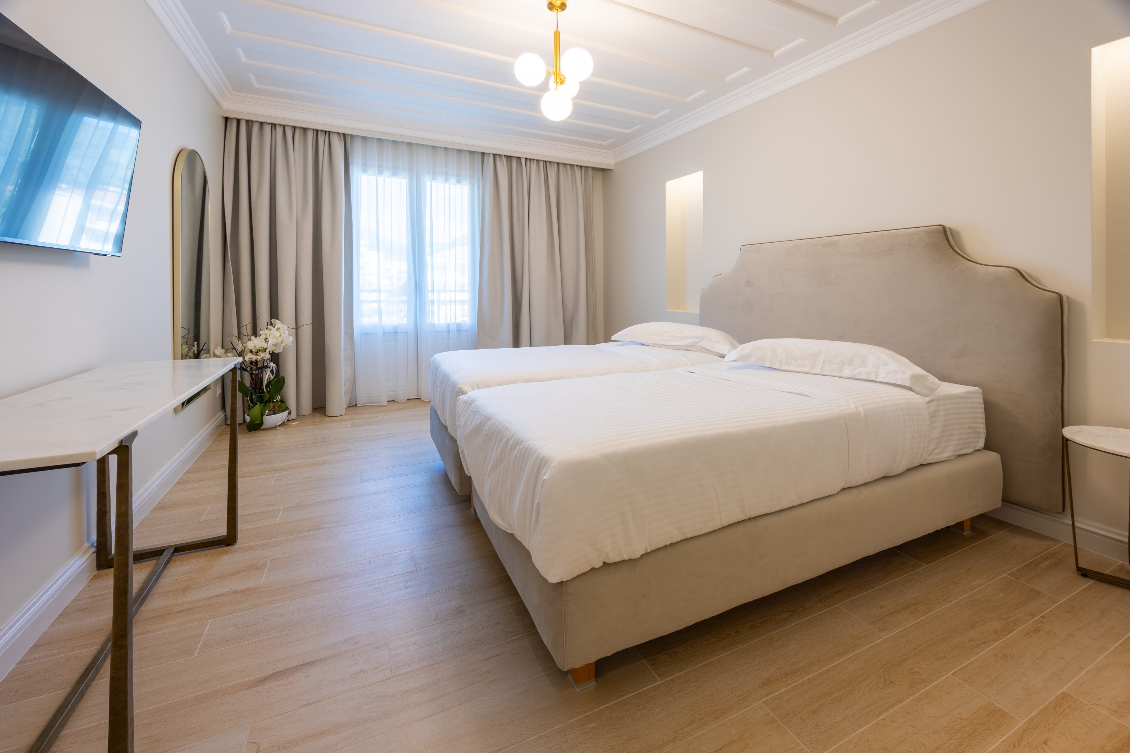 A contemporary hotel bedroom features two neatly made twin beds with light upholstered frames. The room is decorated in neutral tones with wooden flooring and soft beige walls. A large window with sheer and blackout curtains lets in natural light, while a ceiling light fixture and wall niches add warm illumination. A wall-mounted television, slim console table, and decorative mirror complete the minimalist, elegant interior.