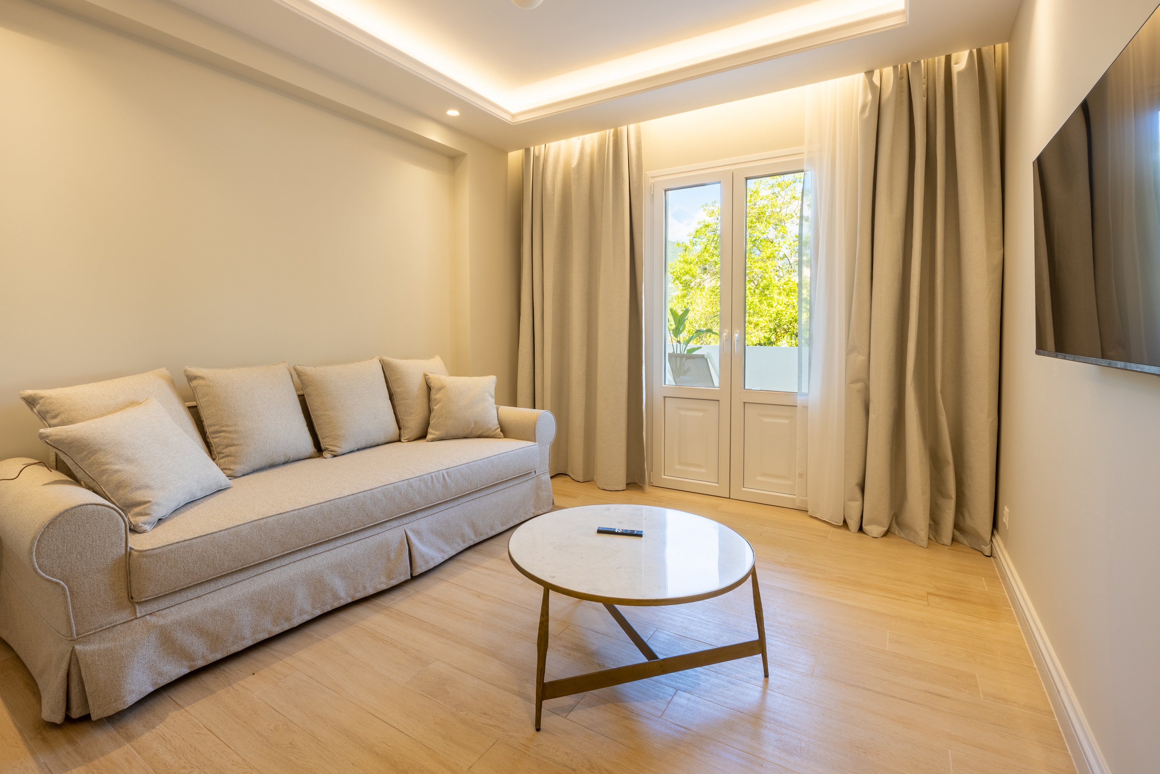 A modern living room features a light-colored sofa with multiple cushions against a neutral wall. A round coffee table with a remote control sits on a light wooden floor. Floor-to-ceiling curtains frame glass doors that open to a view of green trees outside. Recessed ceiling lighting and a wall-mounted television complete the clean, minimalist interior design.