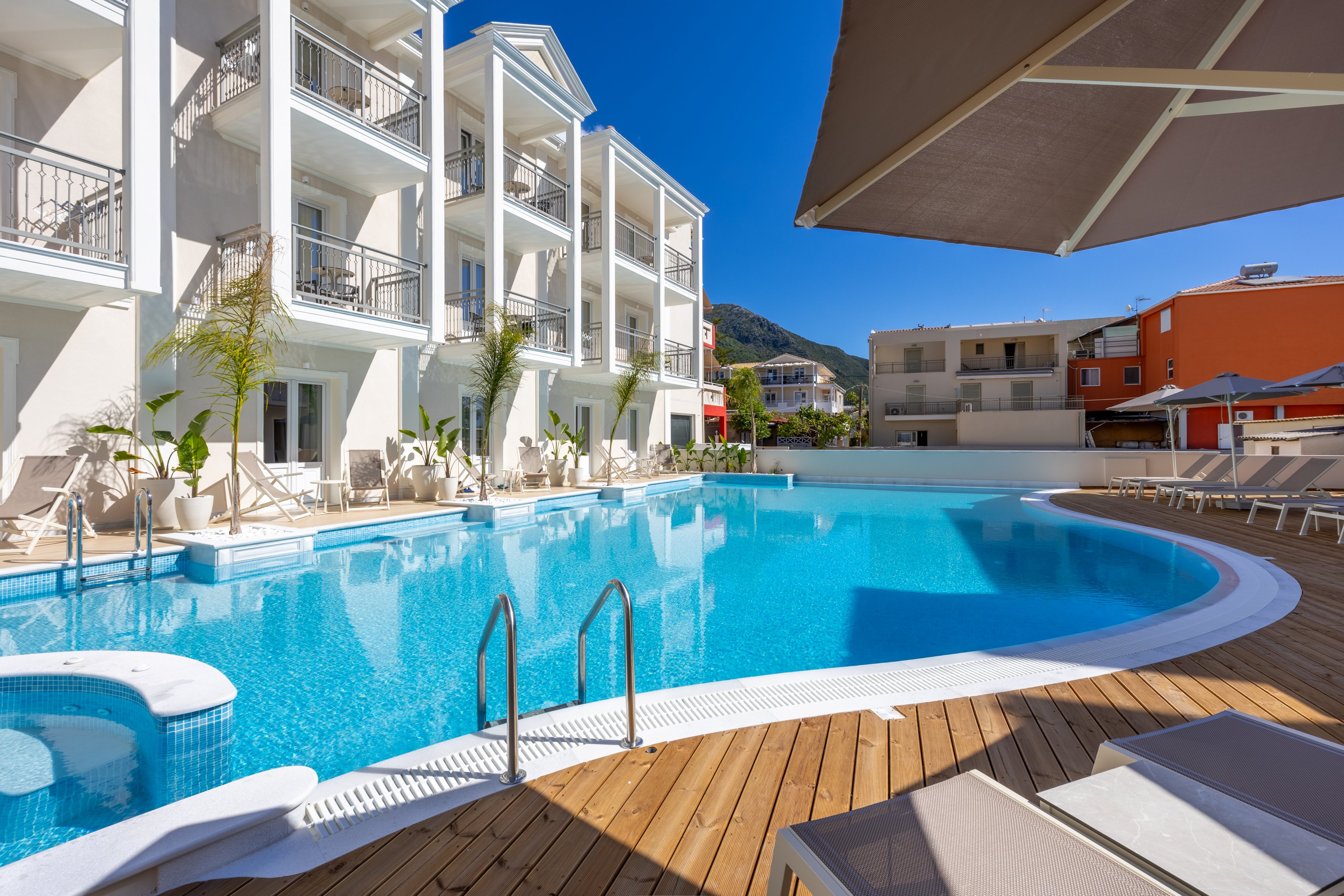 A large curved swimming pool sits in front of a modern multi-story hotel building. Sun loungers and umbrellas line the wooden deck around the clear blue water. Nearby residential and hotel buildings are visible in the background under a bright blue sky. The scene suggests a relaxing vacation atmosphere in a warm, sunny destination.