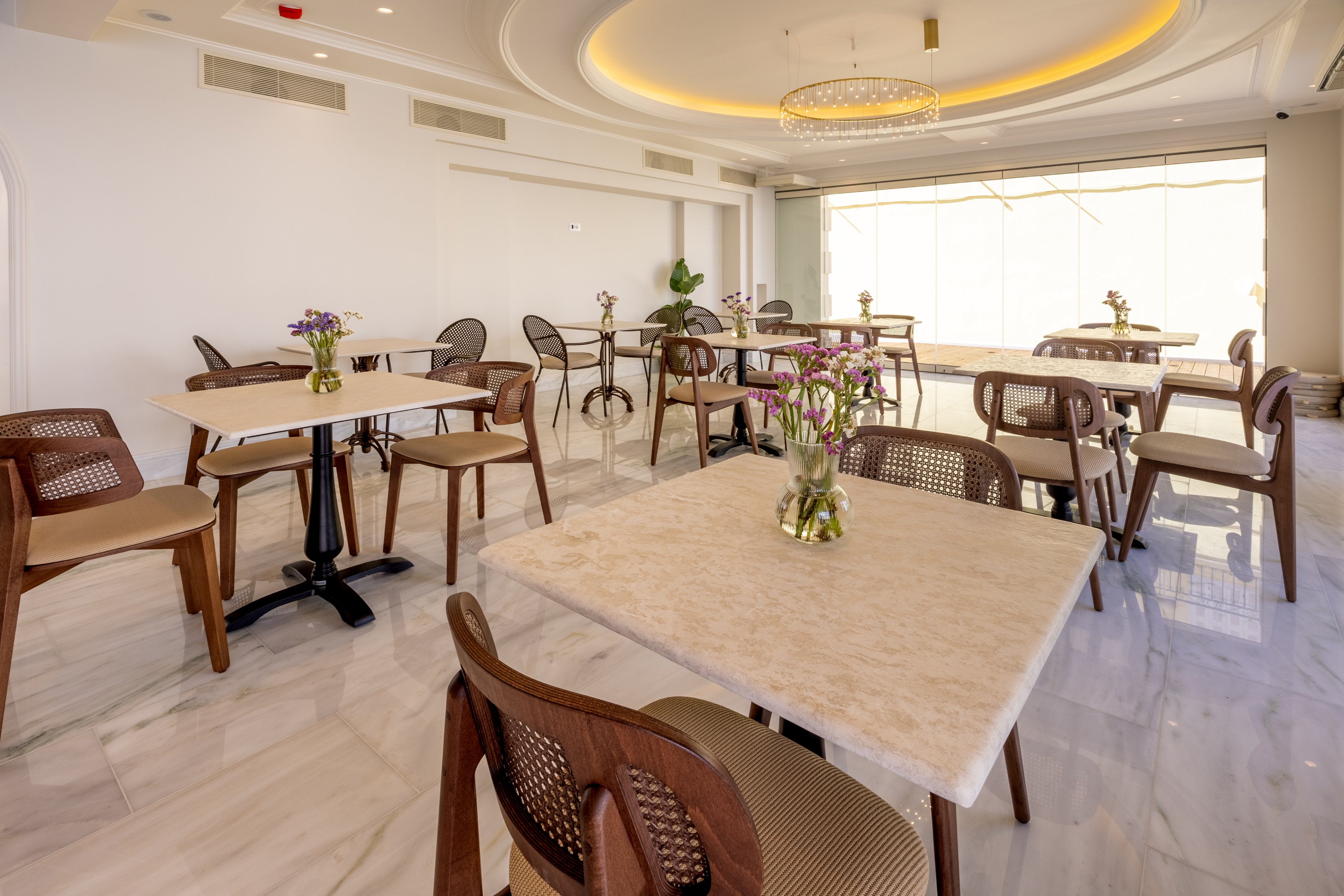 A contemporary cafe dining area features several square tables with light stone tops and wooden chairs. Small vases with fresh flowers decorate the tables, adding a touch of color to the neutral interior. Large windows with sheer coverings allow natural light to fill the space, highlighting the glossy marble floor. A circular ceiling design with recessed lighting and a chandelier enhances the elegant, airy atmosphere.