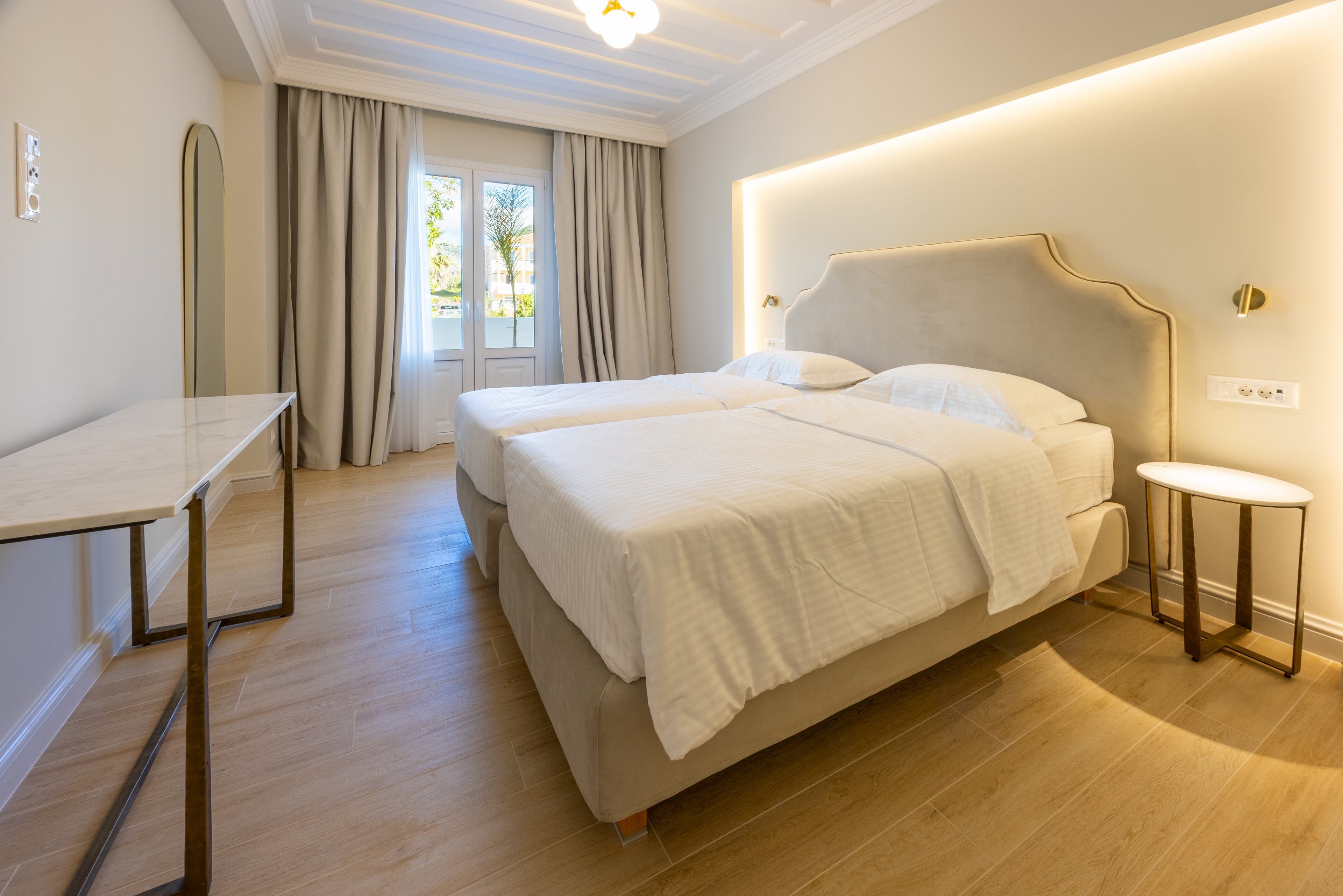 A contemporary hotel room features two neatly made twin beds with white linens and a soft upholstered headboard. The space is bright, with natural light streaming in through glass doors that open to an outdoor view. Warm wooden flooring, neutral walls, and soft curtains create a calm, minimalist atmosphere. A slim desk, tall mirror, and small round bedside table complete the clean, modern interior design.
