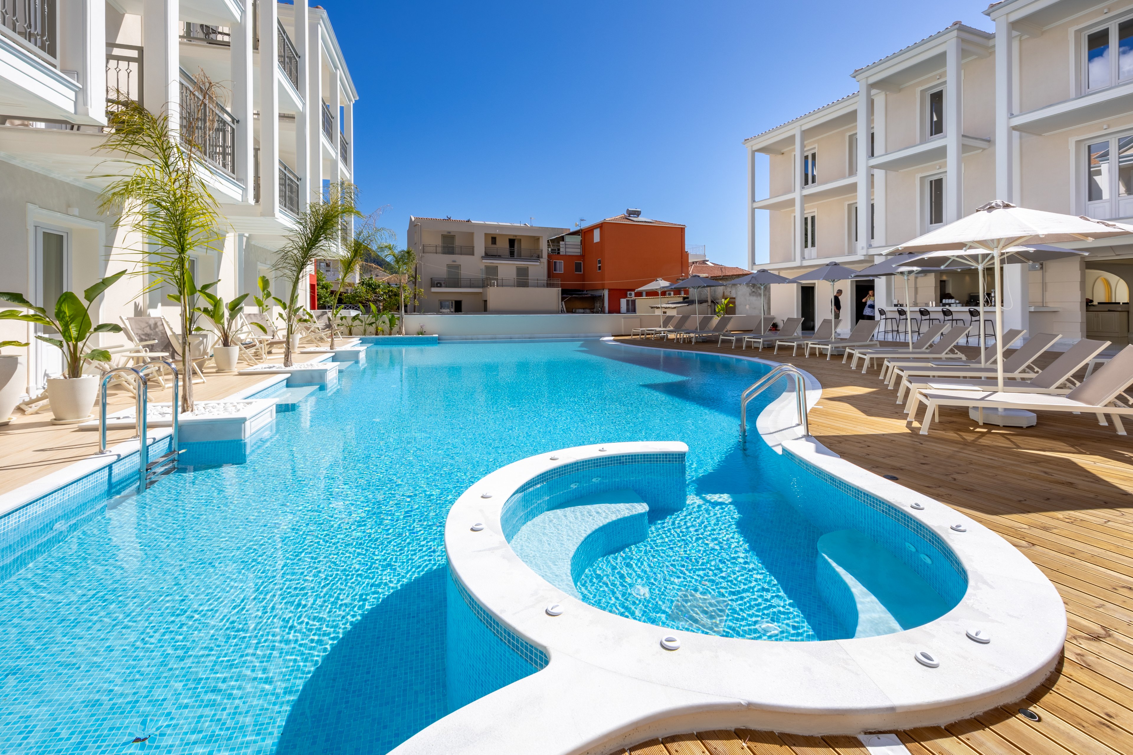 A large, curved swimming pool is surrounded by modern white resort buildings under a clear blue sky. Sun loungers and umbrellas line the wooden deck, creating a relaxing vacation atmosphere. Palm trees add a tropical touch to the bright, clean architecture. The scene is empty of people, emphasizing tranquility and leisure.
