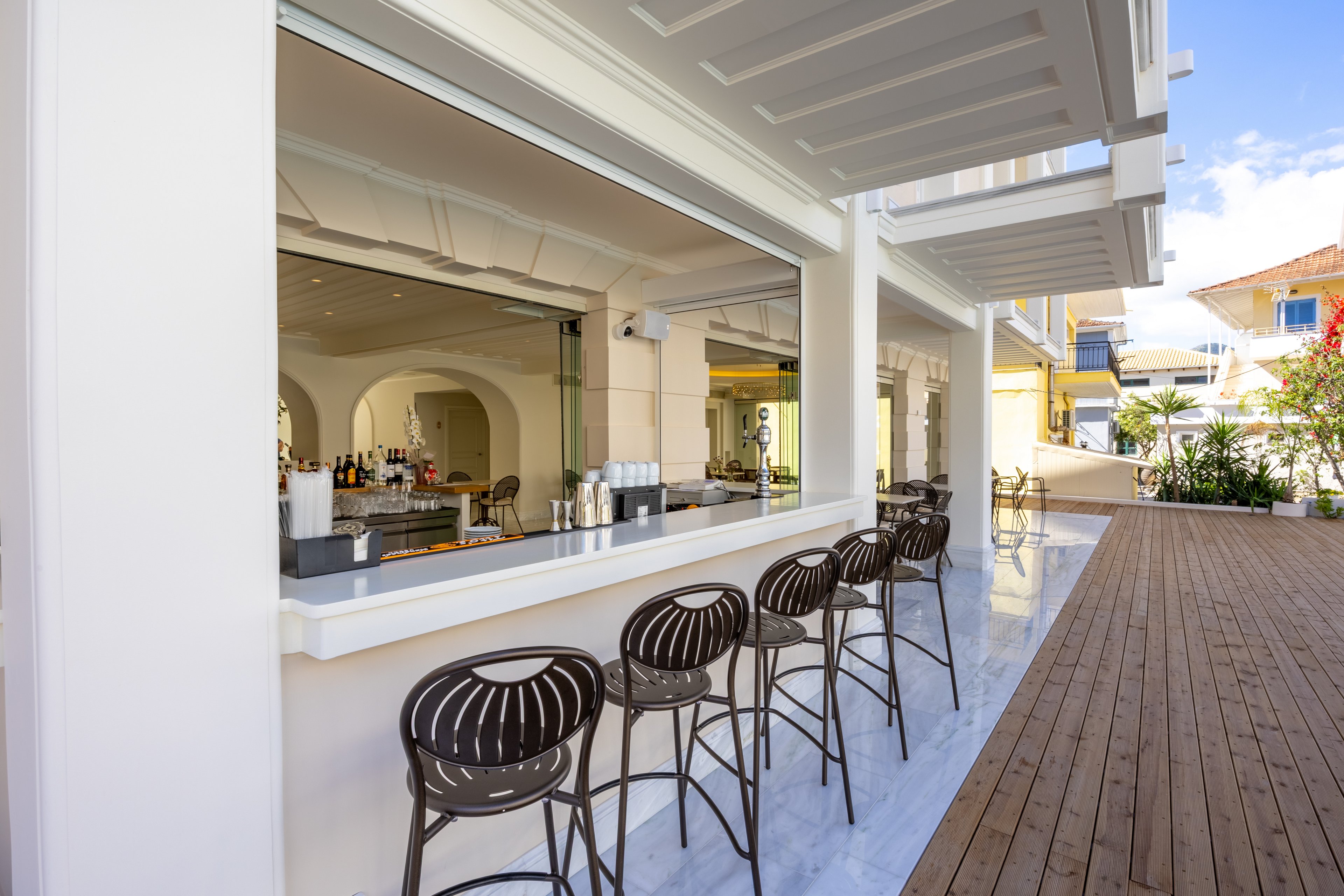 An open-air bar with a sleek white counter features several modern metal bar stools neatly lined up. The bar opens onto a wooden deck walkway under a covered terrace, suggesting a resort or hotel setting. Bottles and glassware are visible inside the bar area, while bright daylight and blue sky create a relaxed vacation atmosphere. Architectural details in white and neutral tones emphasize a clean, contemporary design.