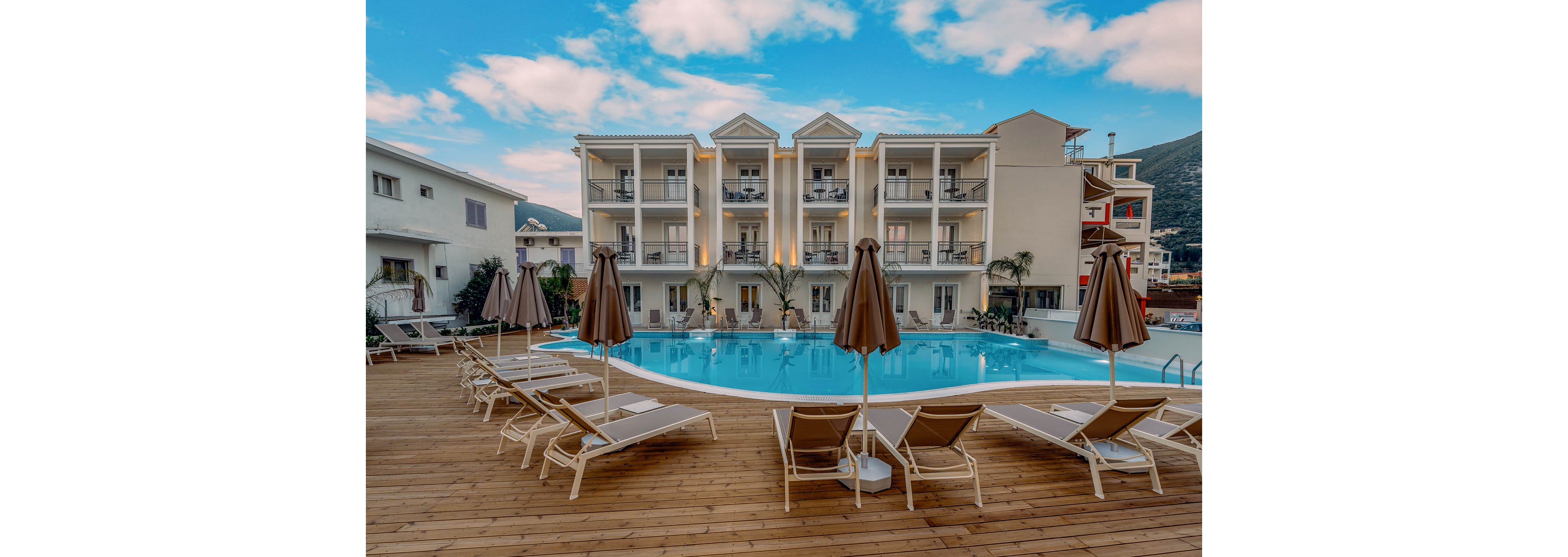 A modern three-story hotel building overlooks a curved outdoor swimming pool. Numerous sun loungers and closed umbrellas are arranged neatly on a wooden deck around the pool. Warm lights from the balconies contrast with the cool blue water and sky, creating a relaxed vacation atmosphere. The scene is calm and empty of people, emphasizing the tranquil resort setting.