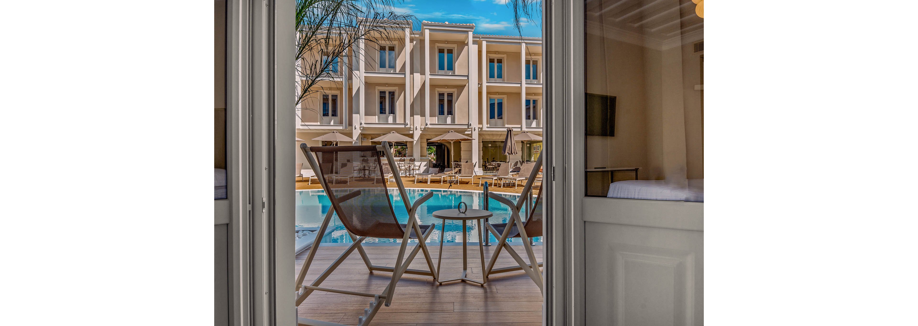 A hotel balcony with two modern lounge chairs and a small round table overlooks a clear blue swimming pool. The scene is framed by open glass doors from an interior guest room, suggesting a relaxing vacation setting. In the background, a multi-story beige hotel building with balconies and poolside seating is visible under a bright blue sky. The overall atmosphere feels calm, sunny, and inviting for leisure or holiday travel.
