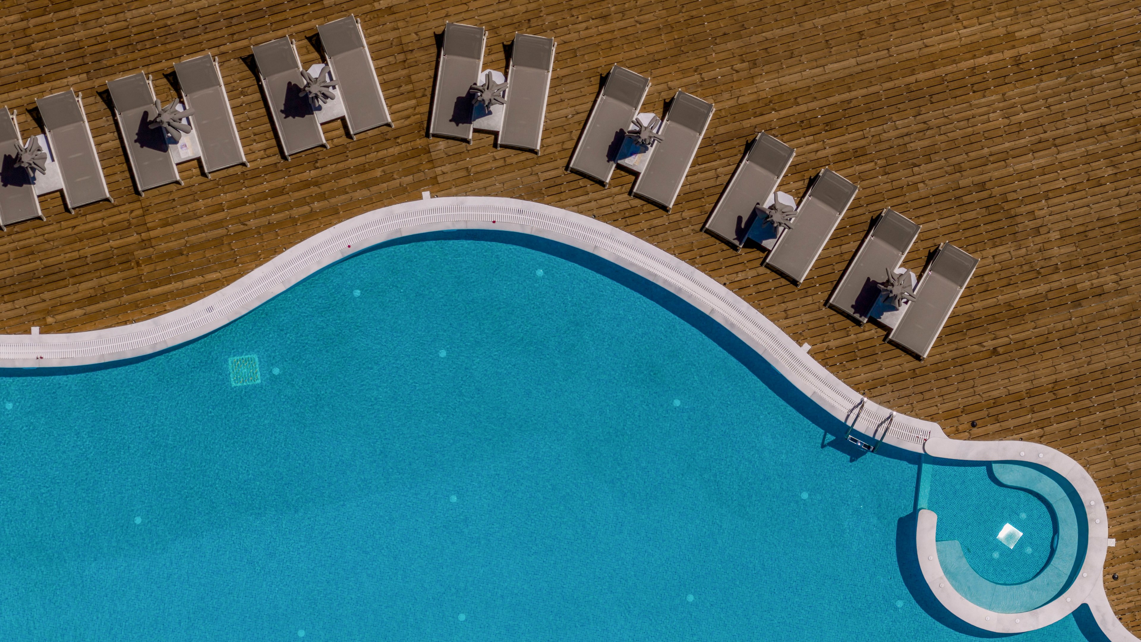 An aerial view shows a large curved swimming pool with clear blue water. A wooden deck lines the pool edge, featuring multiple lounge chairs neatly arranged in rows. A small circular shallow area or spa section is attached to the main pool. The scene suggests a calm, sunny day at a resort or hotel poolside.