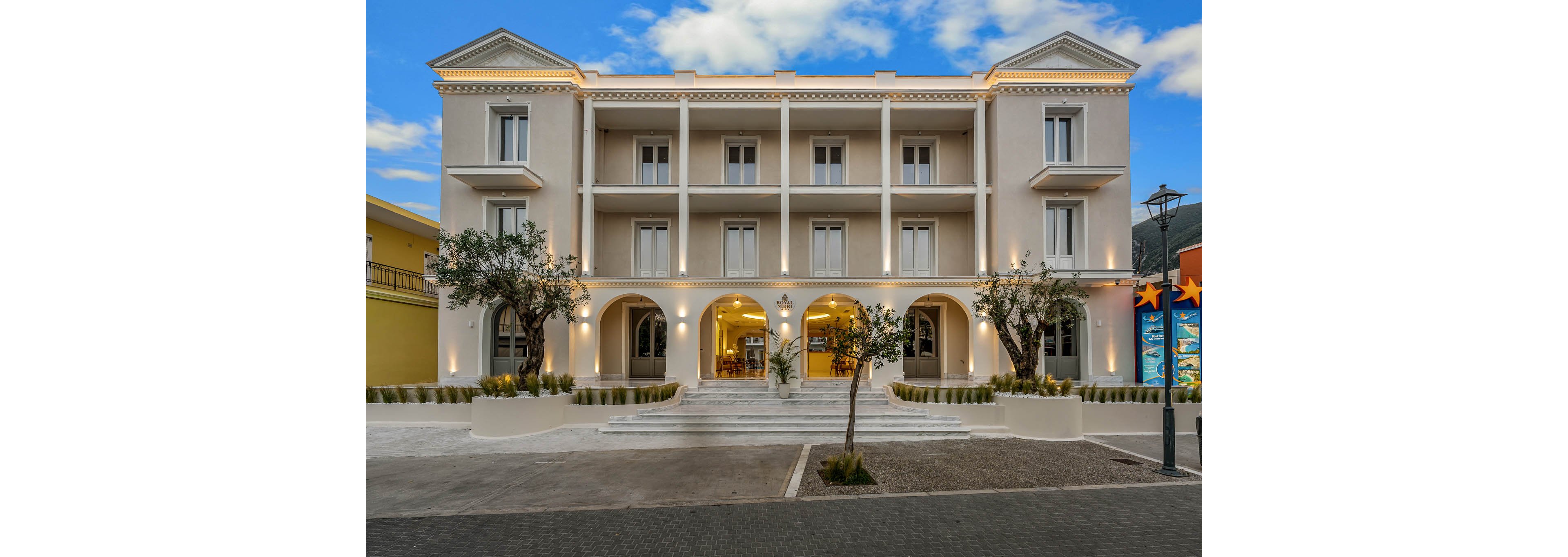 A three-story boutique hotel with a neoclassical facade is photographed at dusk. Warm lights glow from the arched entrance and ground-floor windows, contrasting with the soft beige exterior walls. Two small trees in planters frame the front steps leading up to the main doors. The sky above is bright with scattered clouds, adding a serene atmosphere to the urban streetscape.