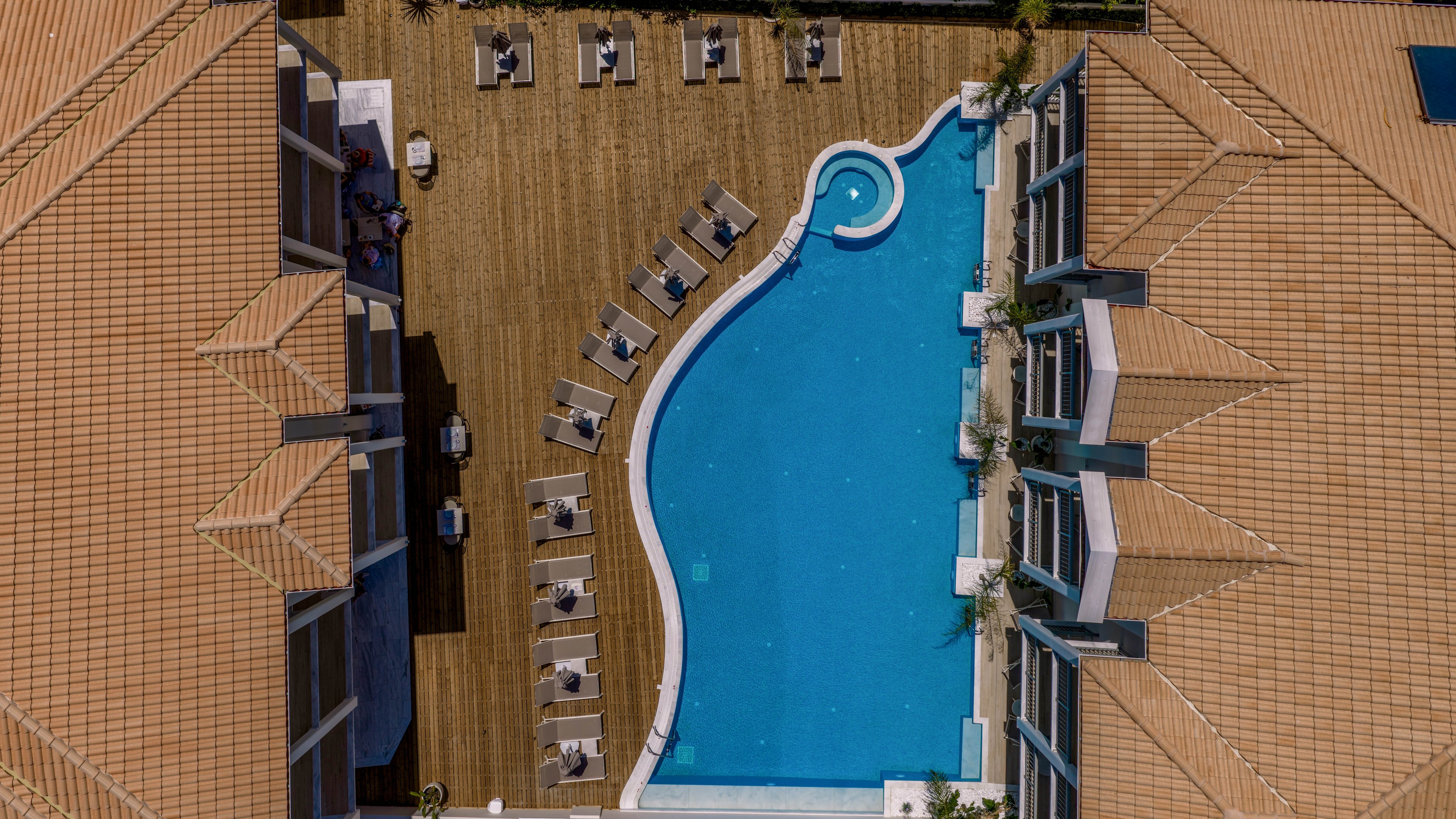An aerial view shows a large, curved swimming pool centered between two tan-roofed buildings. Numerous sun loungers with umbrellas are arranged neatly on the wooden deck surrounding the pool. The clear blue water contrasts with the warm tones of the roofs and decking, creating a clean, modern resort aesthetic. The scene suggests a quiet, sunny day at a contemporary hotel or vacation property.