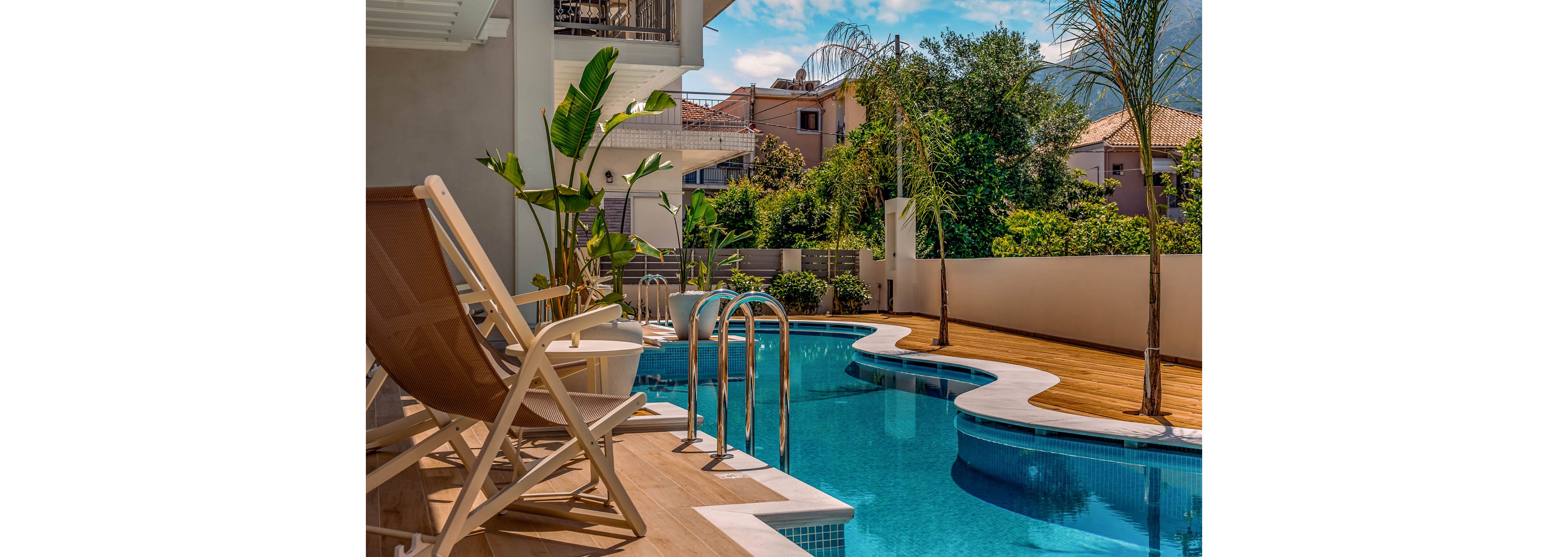A modern residential backyard features a curved swimming pool with clear blue water. Wooden decking surrounds the pool, with lounge chairs and metal ladders providing access and relaxation space. Tropical plants and young palm trees line the edge of the deck, adding a lush, resort-like feel. In the background, neighboring houses and distant mountains sit under a bright, partly cloudy sky.