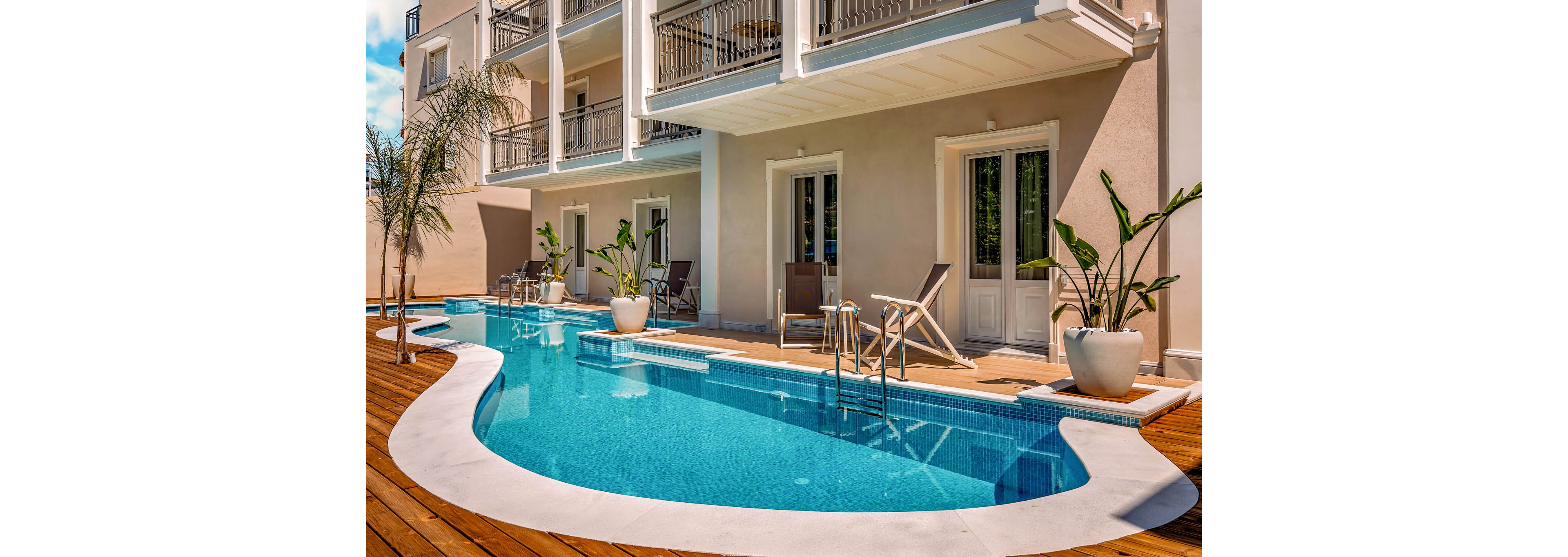 A curved outdoor swimming pool runs alongside a modern hotel or apartment building. White balconies and doors open directly onto the wooden deck, where lounge chairs and a small table with stools are arranged. Potted tropical plants and palm trees add greenery around the bright blue water. The scene is sunlit and tranquil, suggesting a relaxing vacation atmosphere.