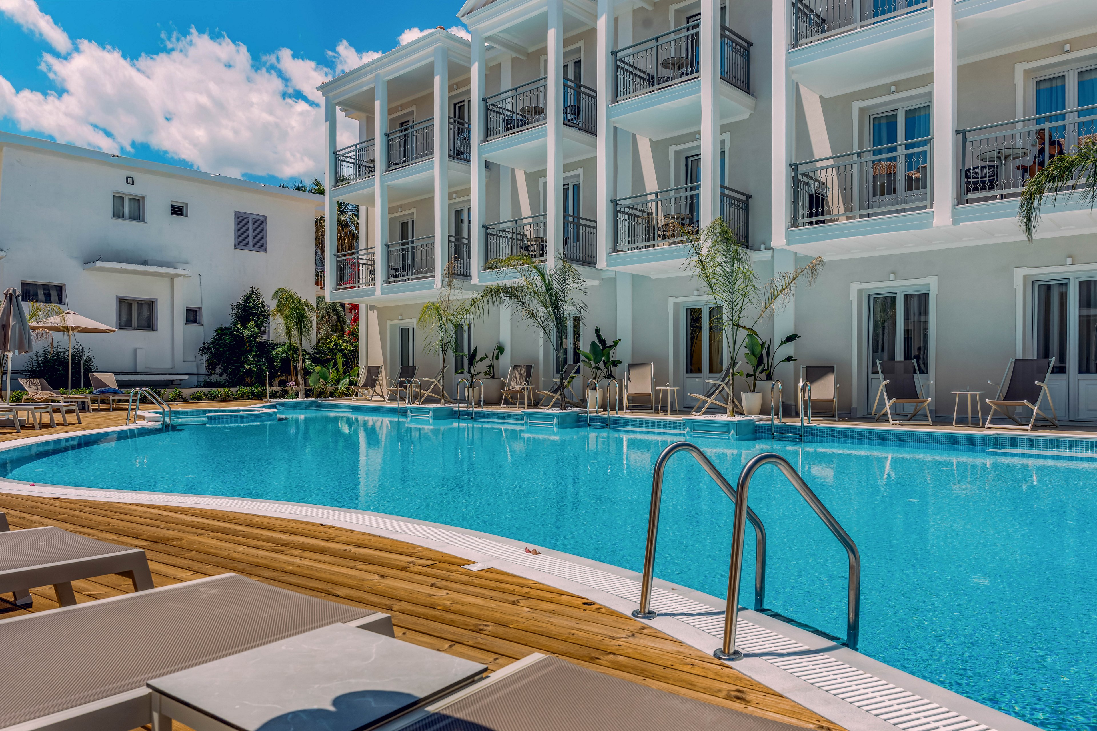 A clear blue swimming pool sits in front of a modern multi-story hotel building. Sun loungers and umbrellas line the wooden deck, creating a relaxed resort atmosphere. The white facade, balconies, and palm trees add to the vacation setting under a bright blue sky with scattered clouds. Stainless steel pool ladders invite guests into the calm water, suggesting a tranquil holiday scene.