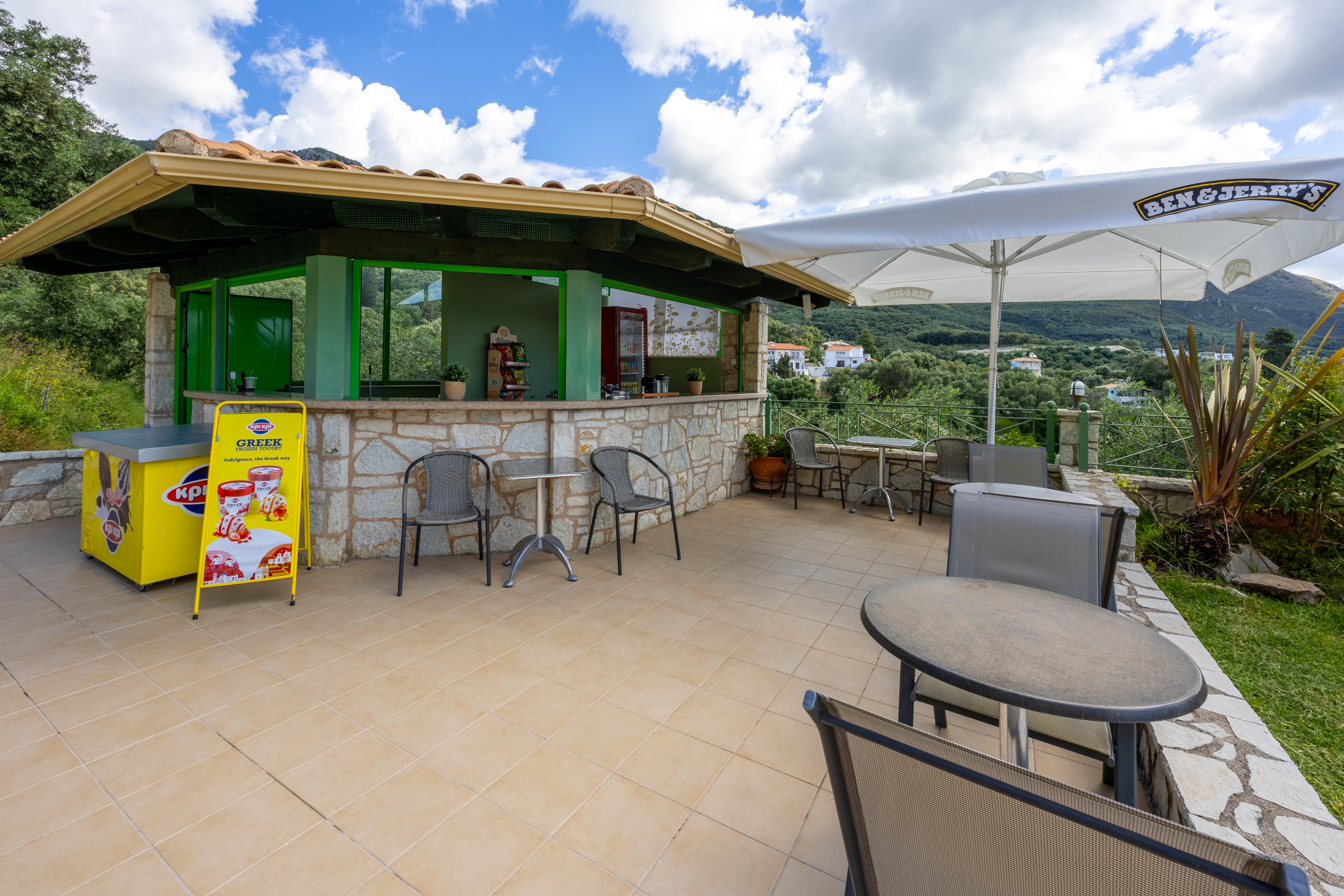 An open-air snack bar with a tiled roof stands on a spacious terrace overlooking green hills. Metal chairs and round tables are arranged on the patio under a large white umbrella. A bright yellow ice cream freezer with colorful product images sits beside the stone counter. The scene is set under a partly cloudy sky, creating a relaxed holiday atmosphere.