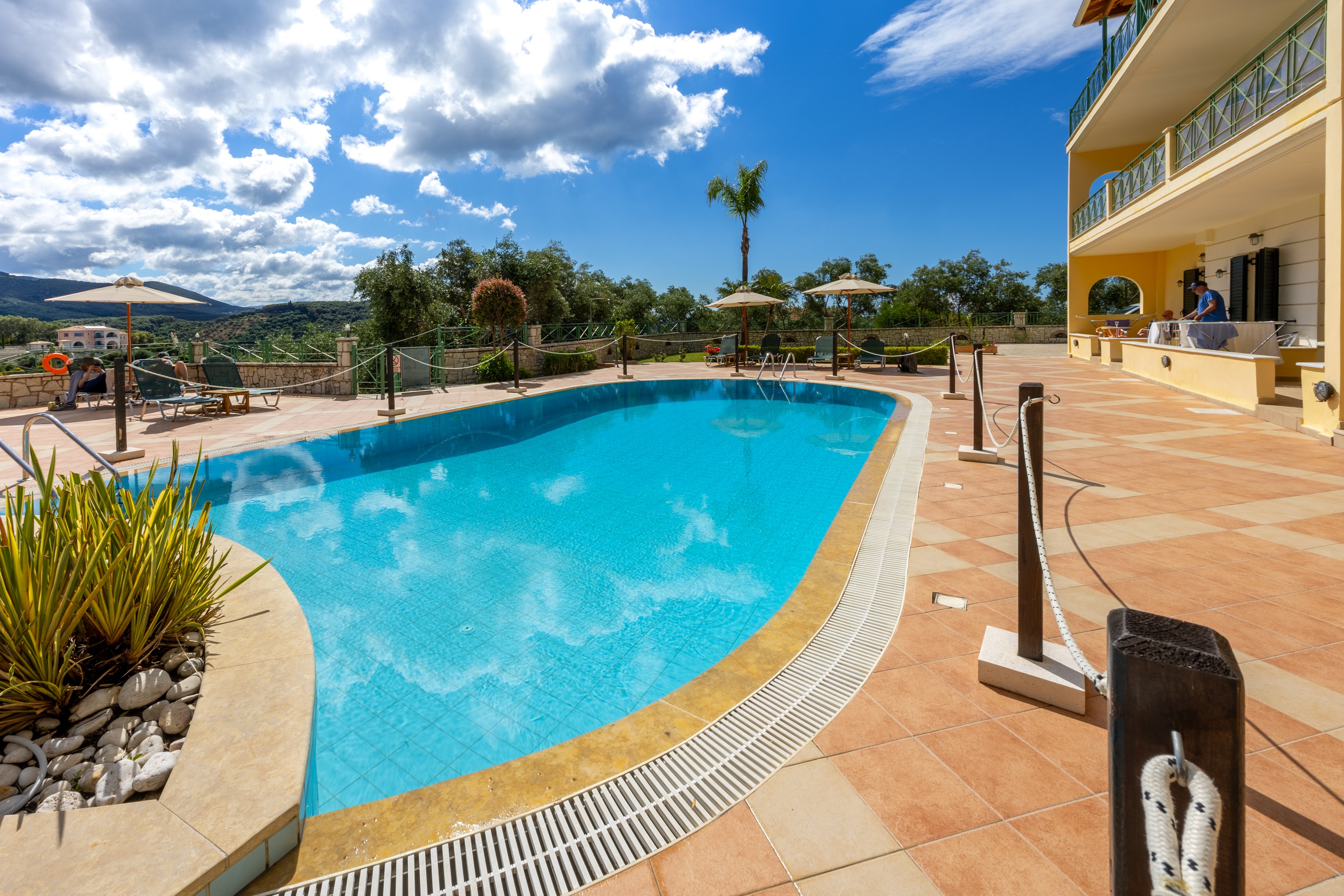 An outdoor swimming pool glistens under bright sunlight beside a resort building. Lounge chairs and umbrellas line the tiled pool deck, inviting guests to relax. Lush greenery and distant hills create a scenic mountain backdrop. A few people stand near the building, adding a subtle sense of activity to the tranquil vacation setting.