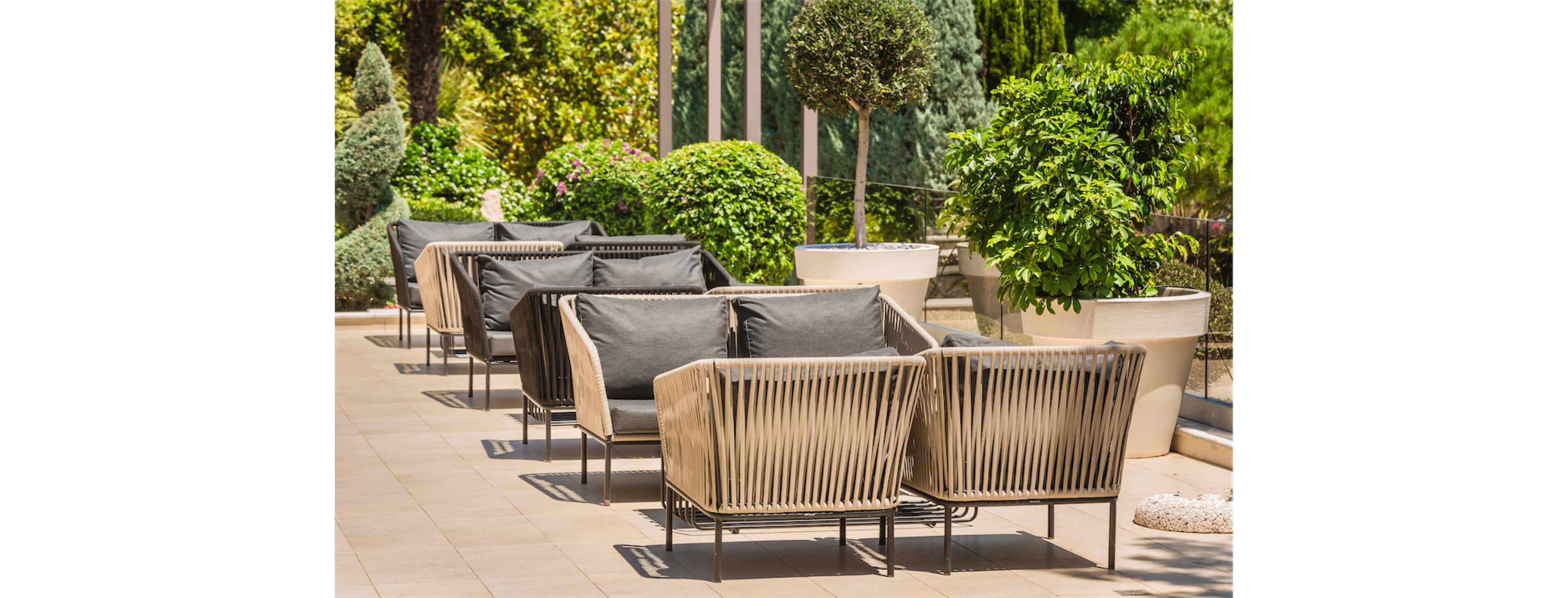 Contemporary outdoor lounge chairs with dark cushions are arranged in pairs along a tiled terrace. The seating features sleek, woven frames and low-profile metal legs. Large potted shrubs and manicured greenery line the edge of the terrace, creating a lush garden backdrop. Bright daylight and clean design elements suggest a relaxed, upscale outdoor living or hospitality space.