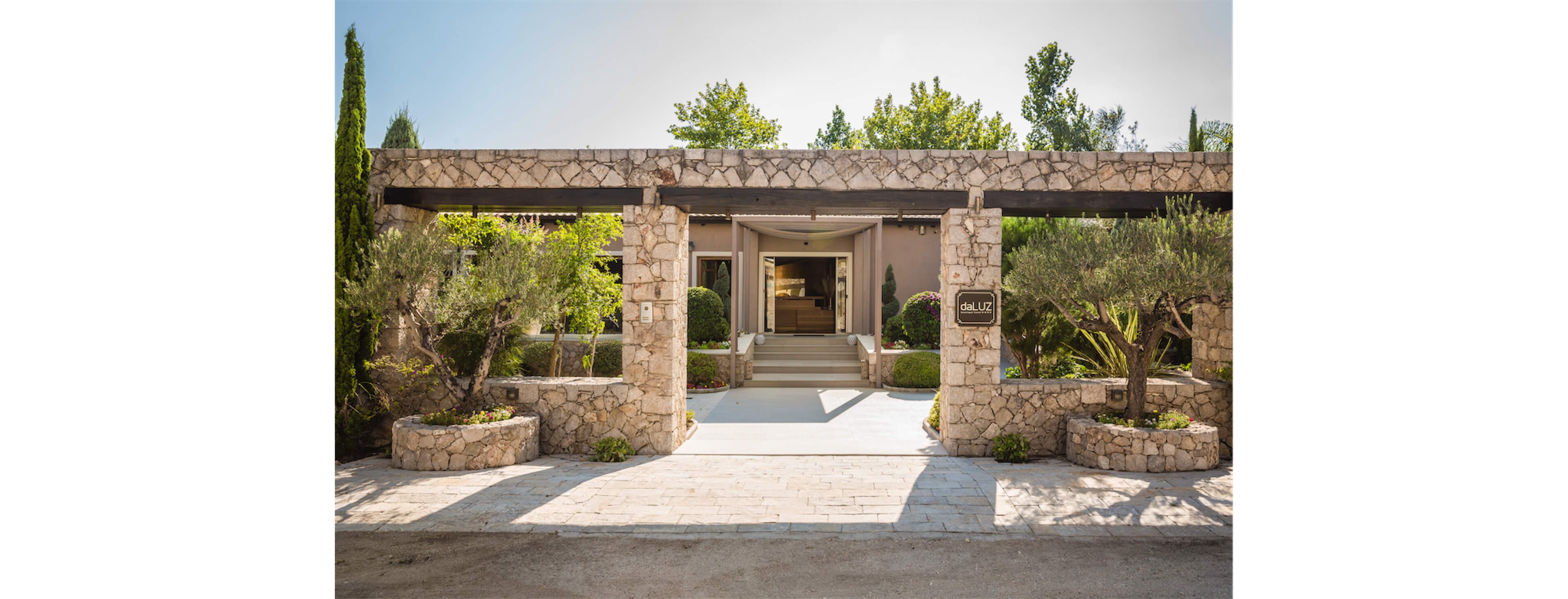 A modern stone villa entrance features a wide pathway leading to a recessed doorway. The facade is built from light-colored stone blocks with rectangular pillars framing the entry. Lush greenery, including trimmed bushes and small trees, softens the architecture and adds a Mediterranean feel. Bright daylight and clear shadows highlight the clean lines and inviting atmosphere of the property.