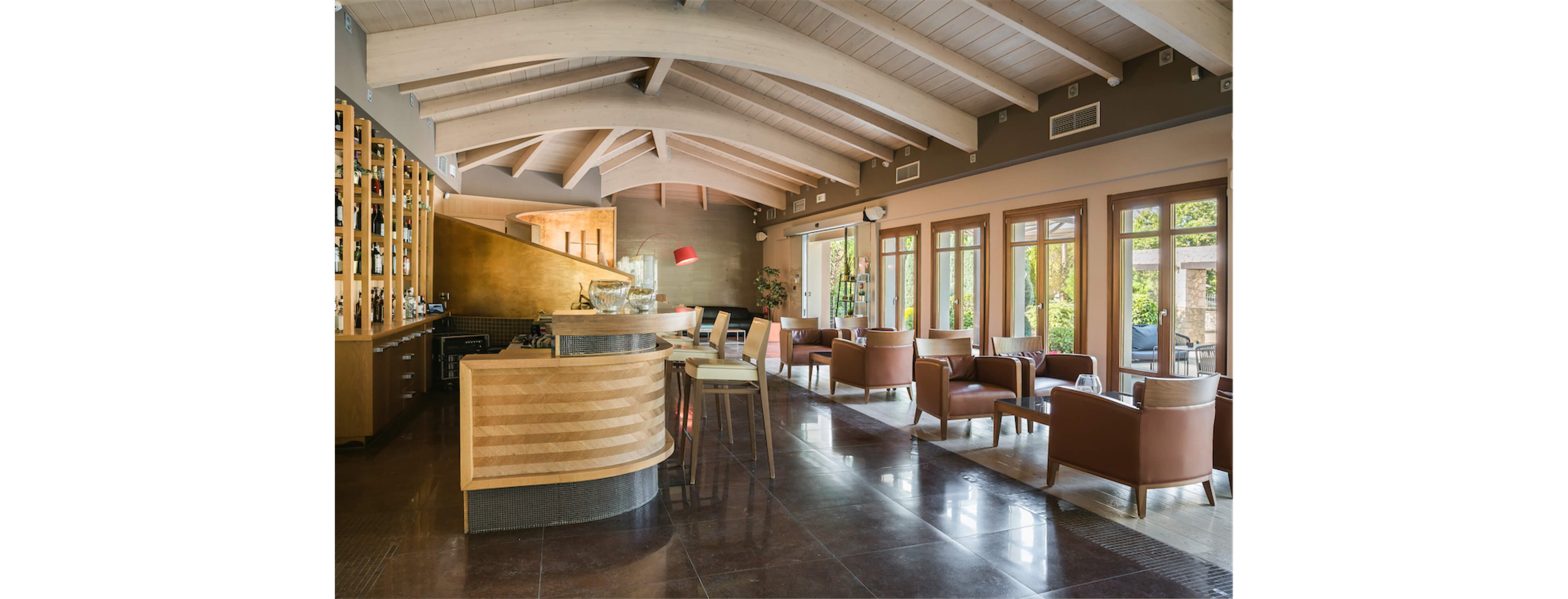 Contemporary lounge bar interior featuring a curved wooden counter and high stools. The space is filled with natural light from large windows and glass doors along one side. Comfortable armchairs and small tables are arranged for relaxed seating throughout the room. Warm neutral tones, wood finishes, and polished floors create an inviting, upscale atmosphere.