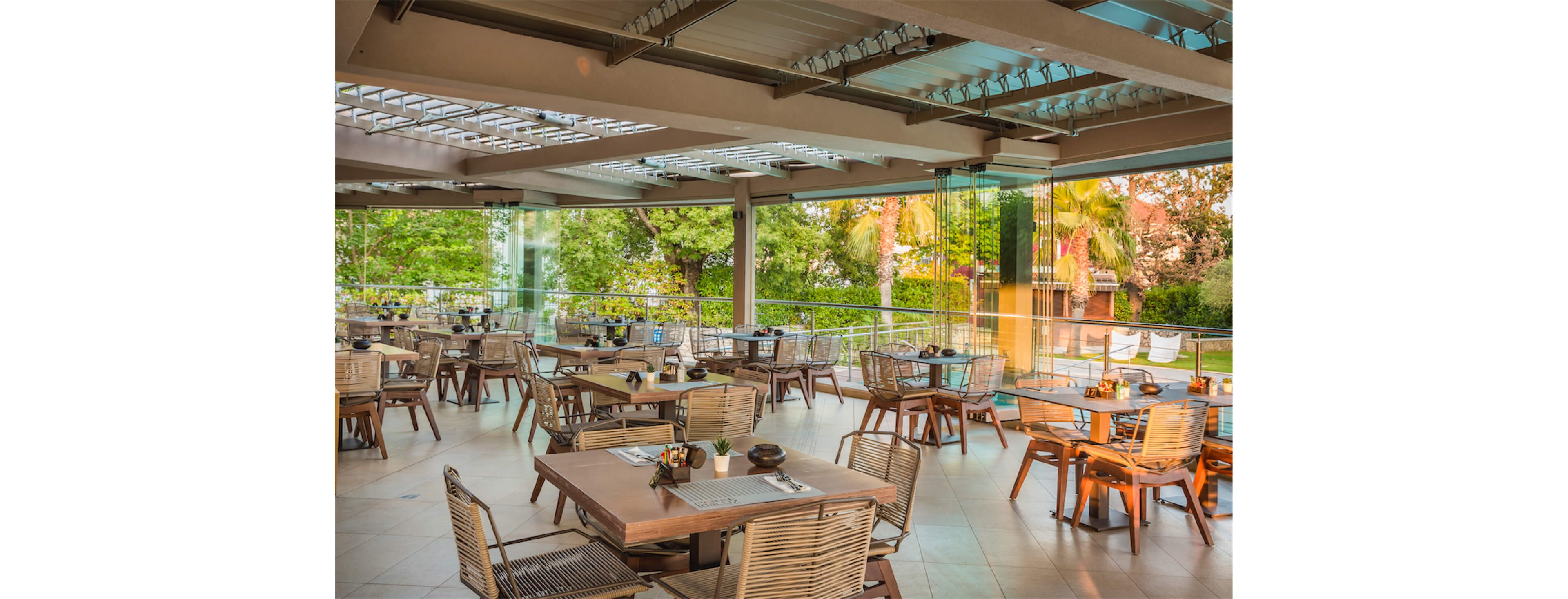 Spacious open-air restaurant terrace featuring neatly arranged wooden tables and chairs. The setting overlooks lush green trees and landscaped gardens through glass railings. Tabletop details include place settings, condiments, and small decorative items, suggesting readiness for guests. Natural light filters through the partially covered roof, creating a relaxed and inviting dining atmosphere.
