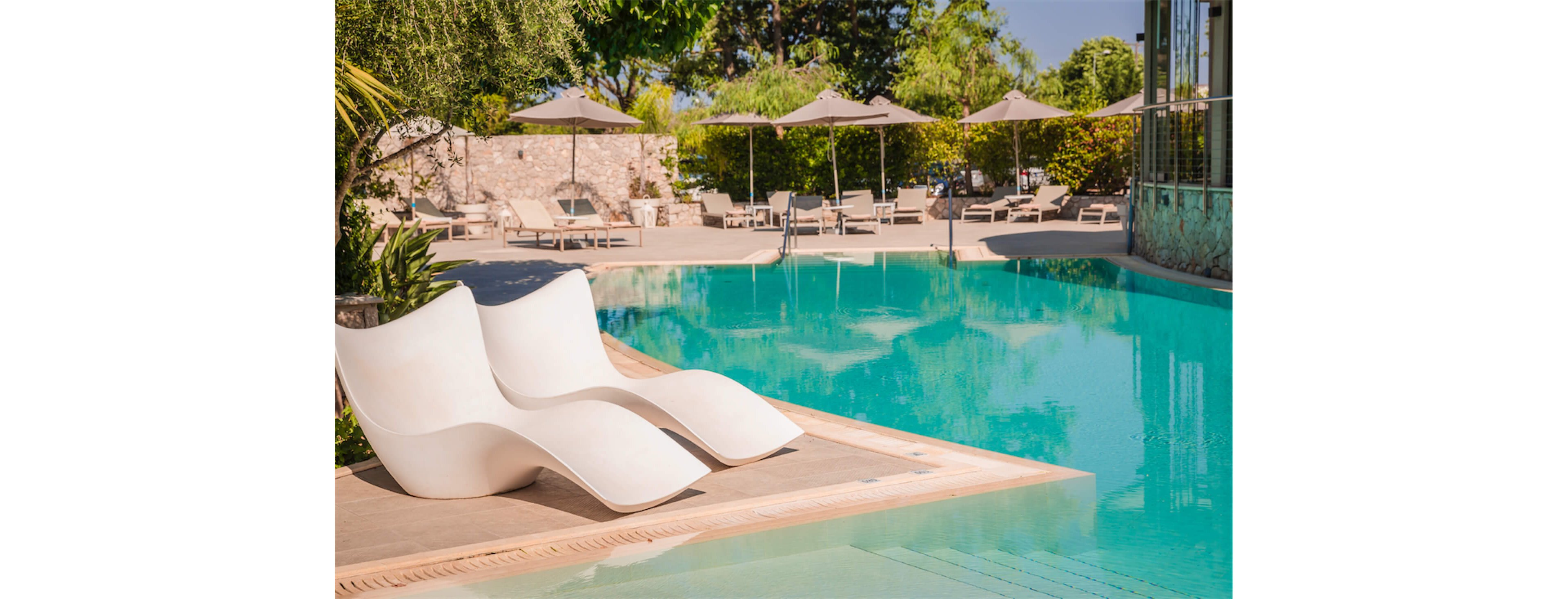 A tranquil outdoor swimming pool features two sculptural white loungers partially submerged at the shallow end. In the background, sunbeds and umbrellas line the pool deck, surrounded by lush greenery and trees. The clear turquoise water reflects the bright daylight, creating a relaxing vacation atmosphere. The scene suggests a modern resort or hotel spa focused on comfort and leisure.